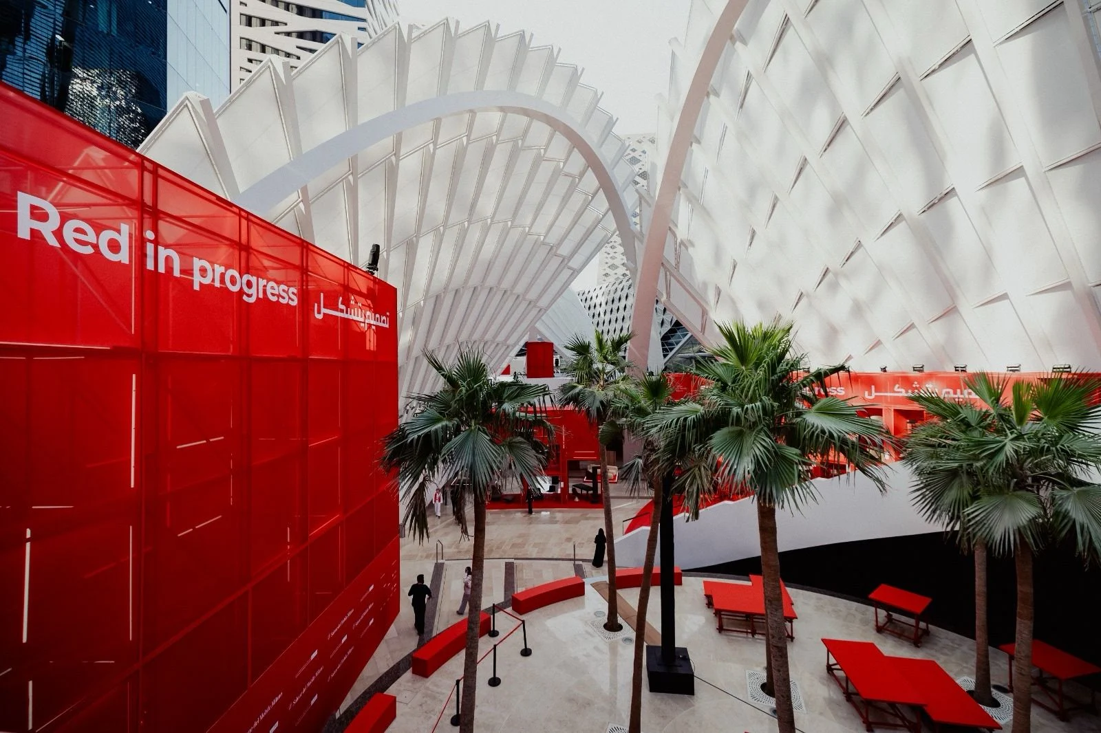 Installazione Red in progress: un volume di vetro rosso e tavoli rossi tra le palme sotto coperture bianche nervate – Red in Progress: Salone del Mobile Meets Riyadh, Arabia Saudita, 2025 – allestimento espositivo