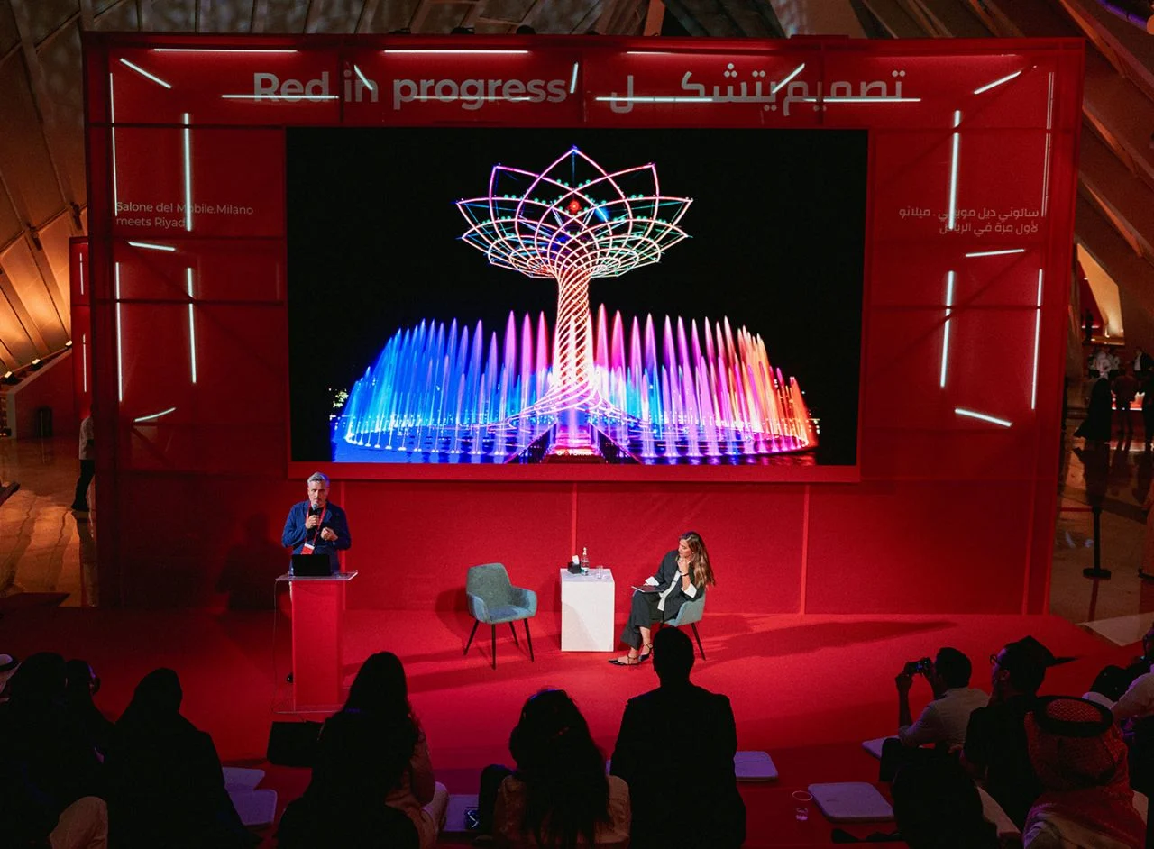 Incontro sul palco rosso, dietro uno schermo con una scultura ad albero illuminata sopra fontane, pubblico davanti – Red in Progress: Salone del Mobile Meets Riyadh, Arabia Saudita, 2025 – allestimento espositivo