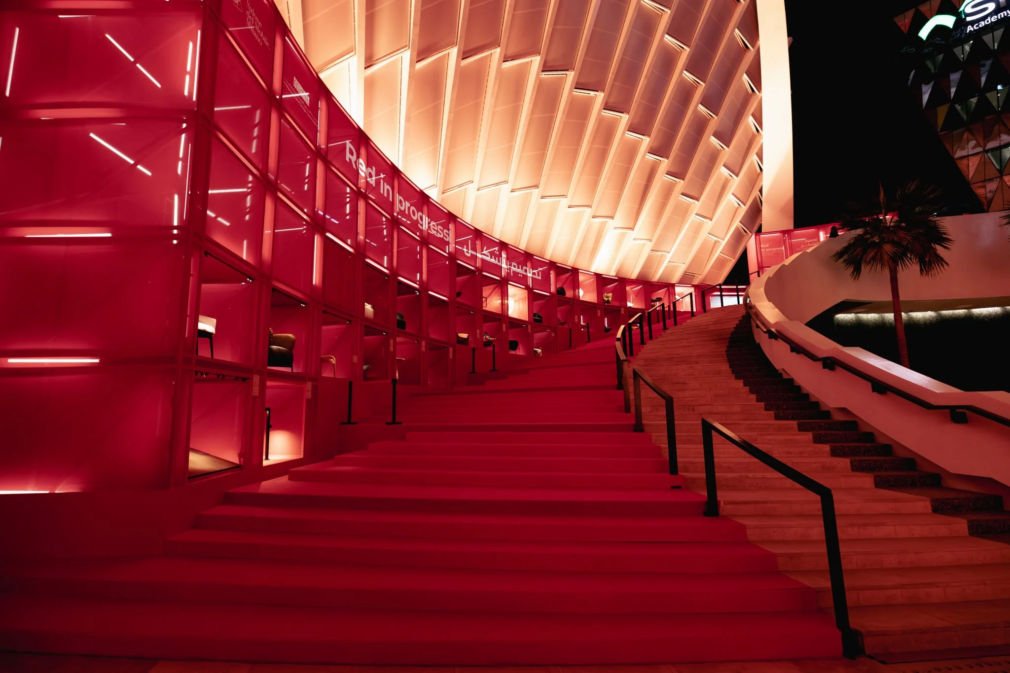 Vista notturna della scala rossa che sale lungo scaffali espositivi illuminati sotto la copertura curva – Red in Progress: Salone del Mobile Meets Riyadh, Arabia Saudita, 2025 – allestimento espositivo