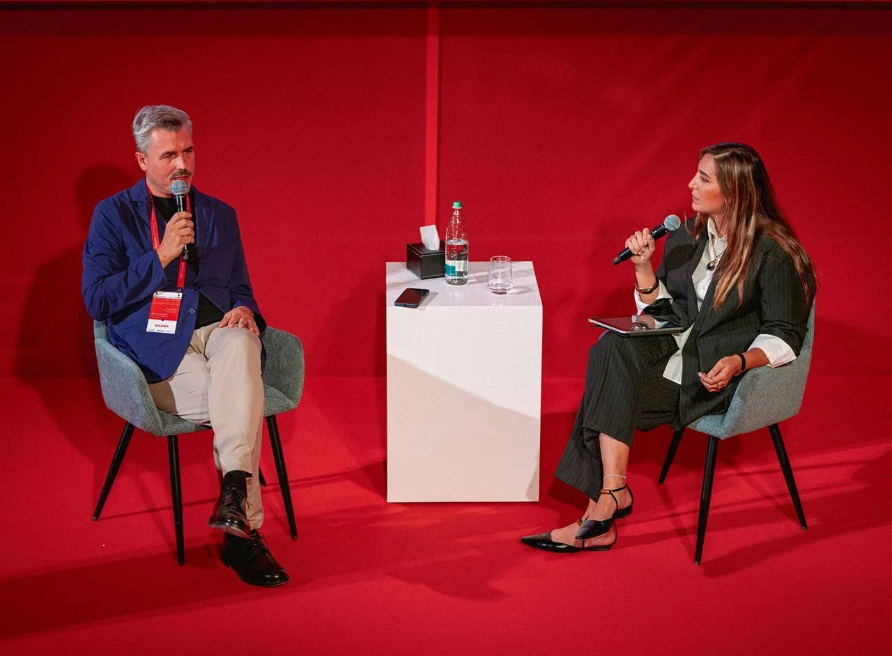 Due relatori seduti a conversare con i microfoni su un palco rosso, un basamento bianco tra loro – Red in Progress: Salone del Mobile Meets Riyadh, Arabia Saudita, 2025 – allestimento espositivo