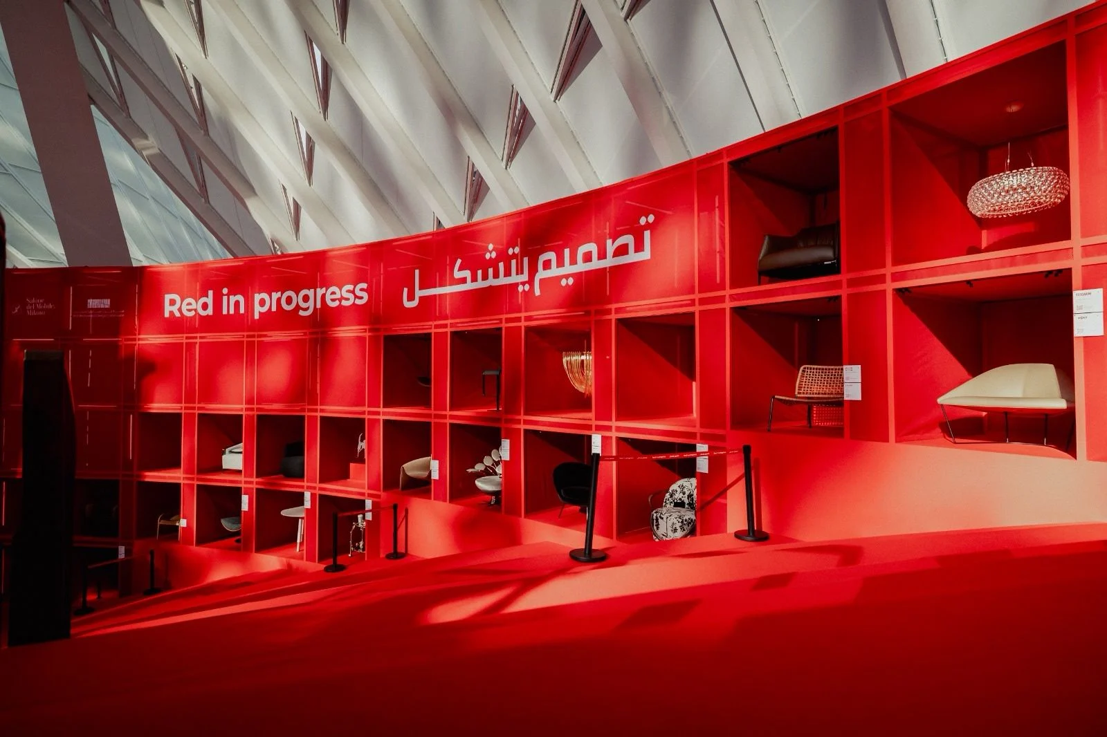 Parete espositiva rossa curva con la scritta Red in progress in inglese e arabo, sedie e lampade nelle nicchie – Red in Progress: Salone del Mobile Meets Riyadh, Arabia Saudita, 2025 – allestimento espositivo
