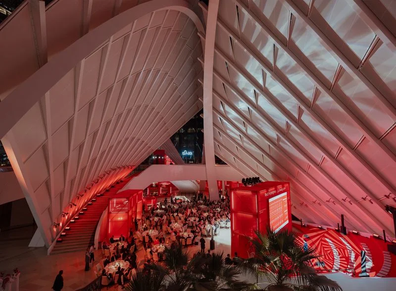 Tavoli della cena di gala sotto la copertura nervata illuminata di rosso, visti dall'alto accanto a una scala rossa – Red in Progress: Salone del Mobile Meets Riyadh, Arabia Saudita, 2025 – allestimento espositivo