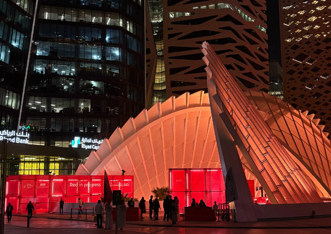 Vista notturna della copertura bianca illuminata e dell'installazione rossa tra le torri per uffici, visitatori nella piazza – Red in Progress: Salone del Mobile Meets Riyadh, Arabia Saudita, 2025 – allestimento espositivo