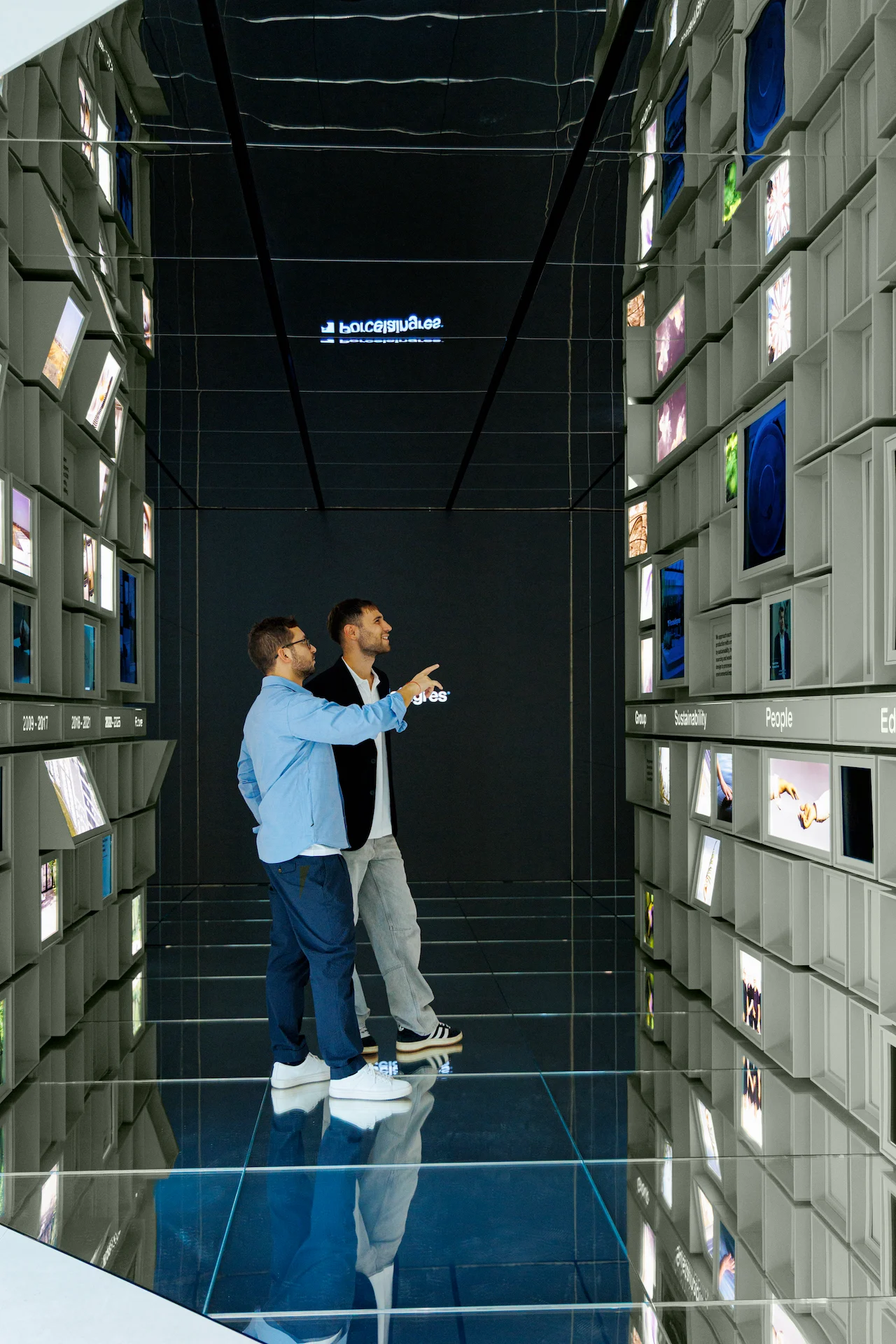Due visitatori nel corridoio a specchi tra pareti di cubi, uno indica uno schermo – Porcelaingres Brand Narrative Space, Castellarano, Italia, 2025 – progettazione di showroom