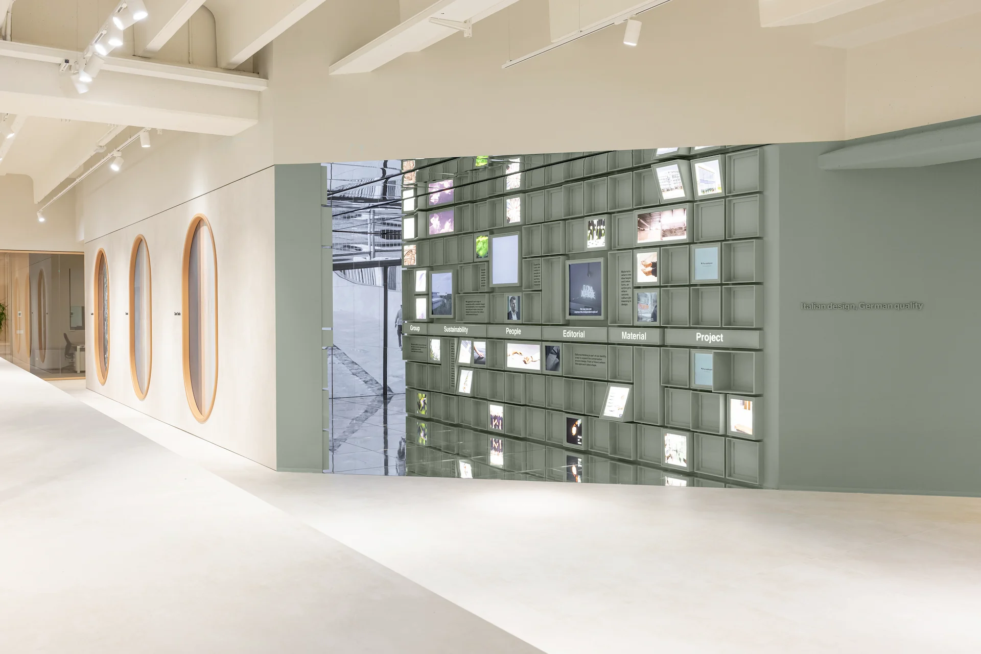 Ingresso dello showroom nello spazio a specchi del marchio: una parete modulare verde salvia di schermi e immagini accanto a nicchie ovali – Porcelaingres Brand Narrative Space, Castellarano, Italia, 2025 – progettazione di showroom