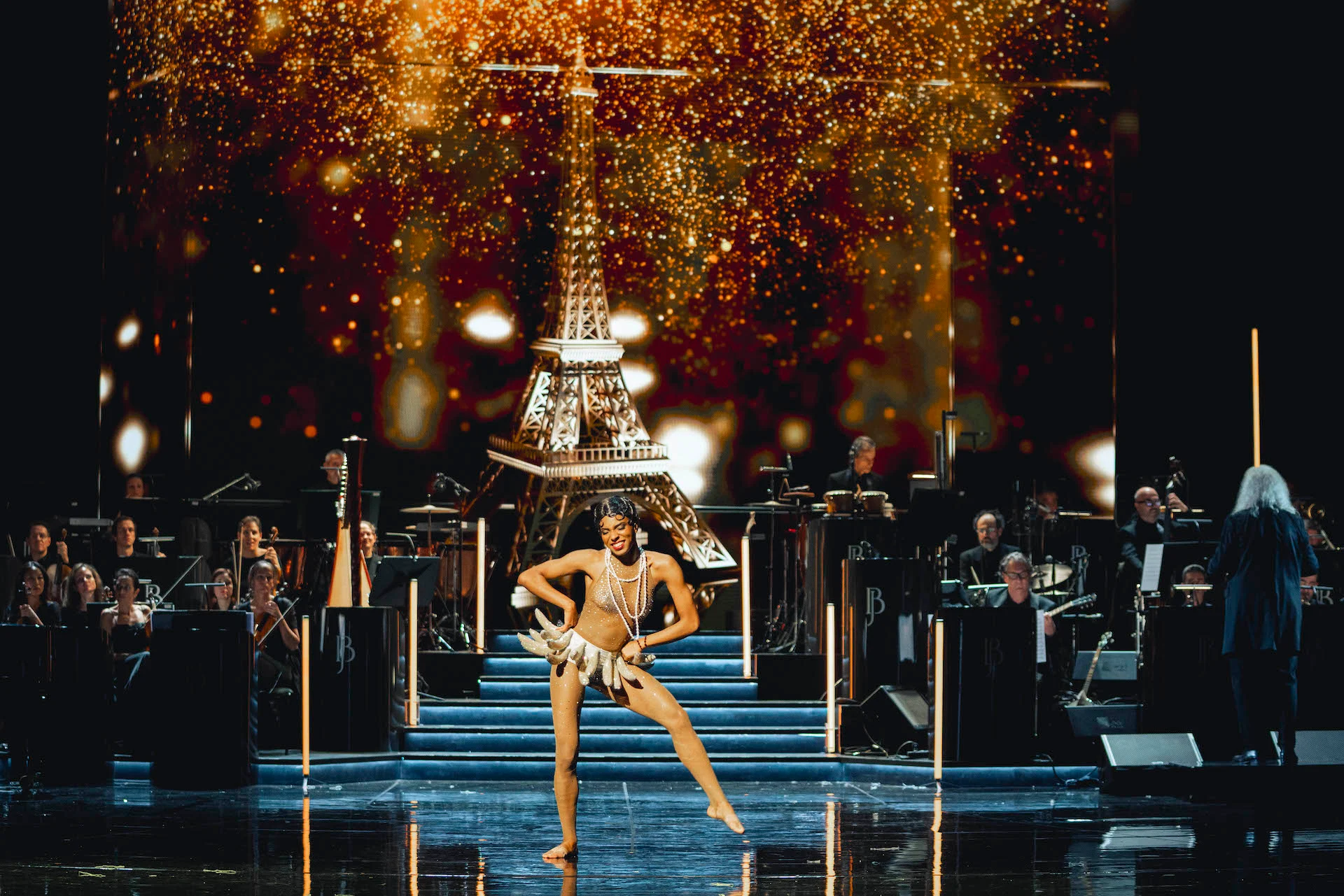 Ballerina con la gonna di banane sui gradini davanti a una Torre Eiffel dorata e luci scintillanti, orchestra dietro – Bonsoir Monte-Carlo, Monte Carlo, Principato di Monaco, 2025 – scenografia
