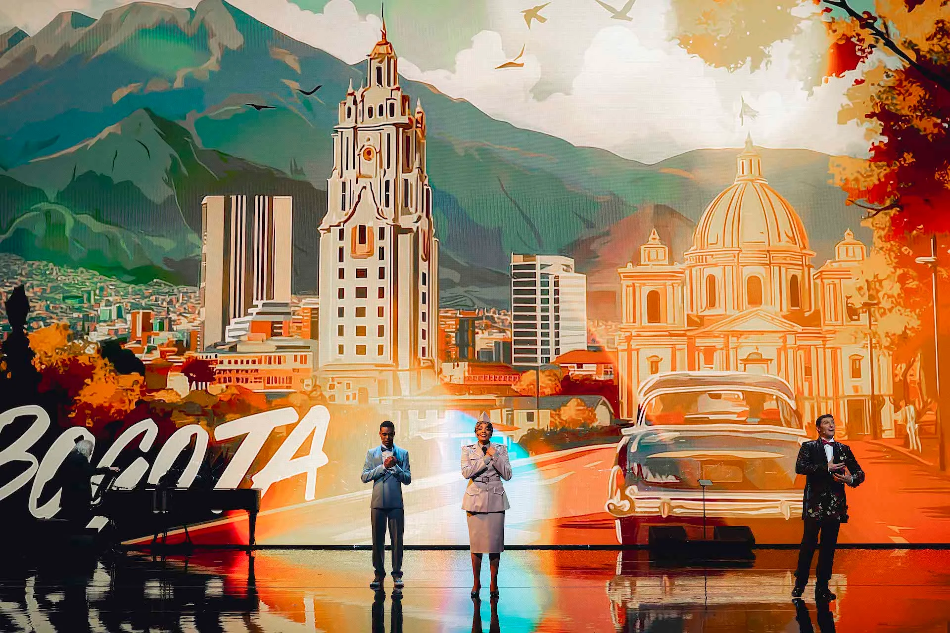 Tre cantanti davanti a un fondale illustrato di Bogotá con montagne, torri, una chiesa con cupola e un'auto d'epoca – Bonsoir Monte-Carlo, Monte Carlo, Principato di Monaco, 2025 – scenografia