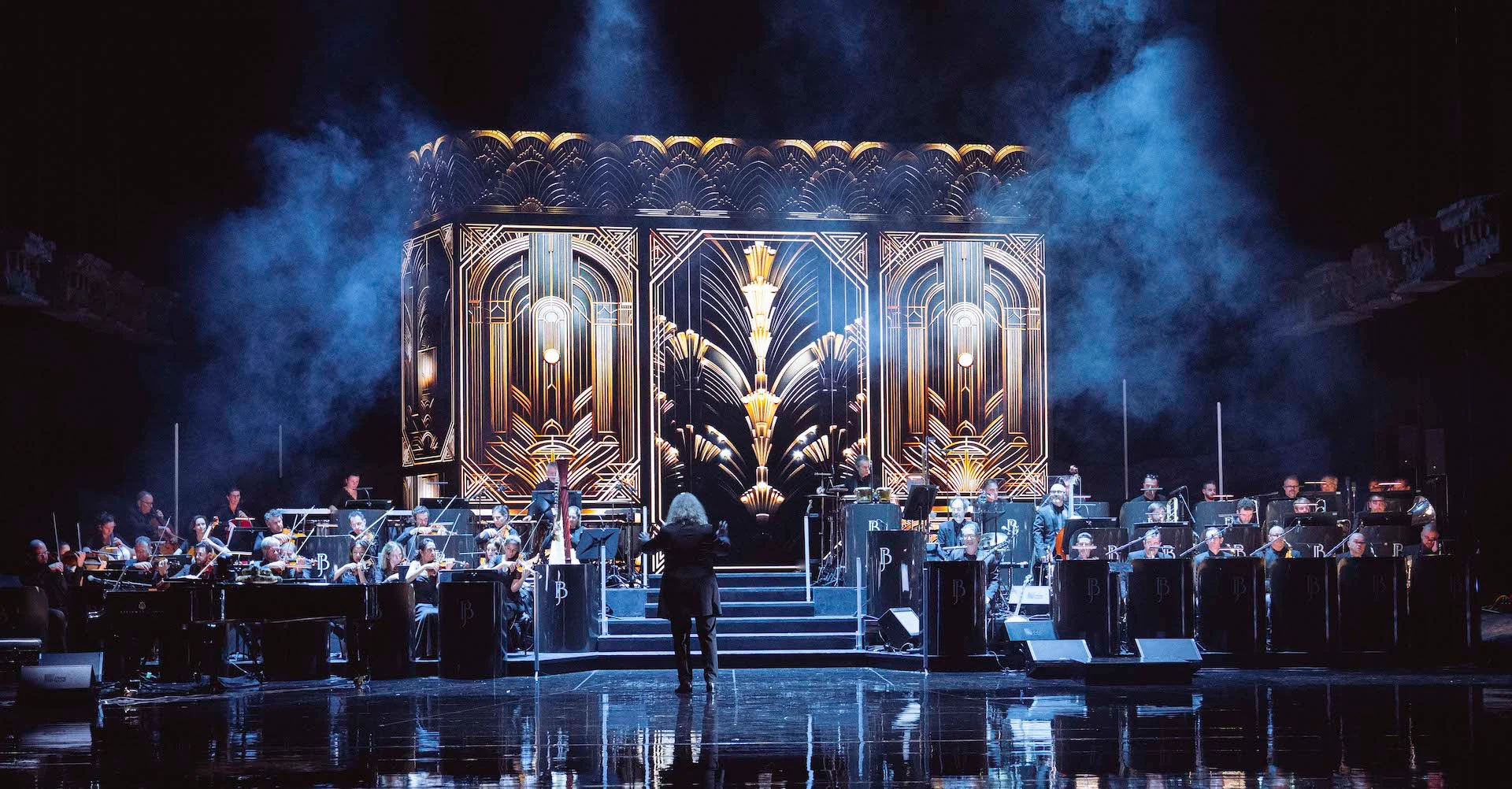 Direttore e orchestra ai lati di una scalinata davanti a un elemento scenico Art Déco oro e nero nella foschia blu – Bonsoir Monte-Carlo, Monte Carlo, Principato di Monaco, 2025 – scenografia
