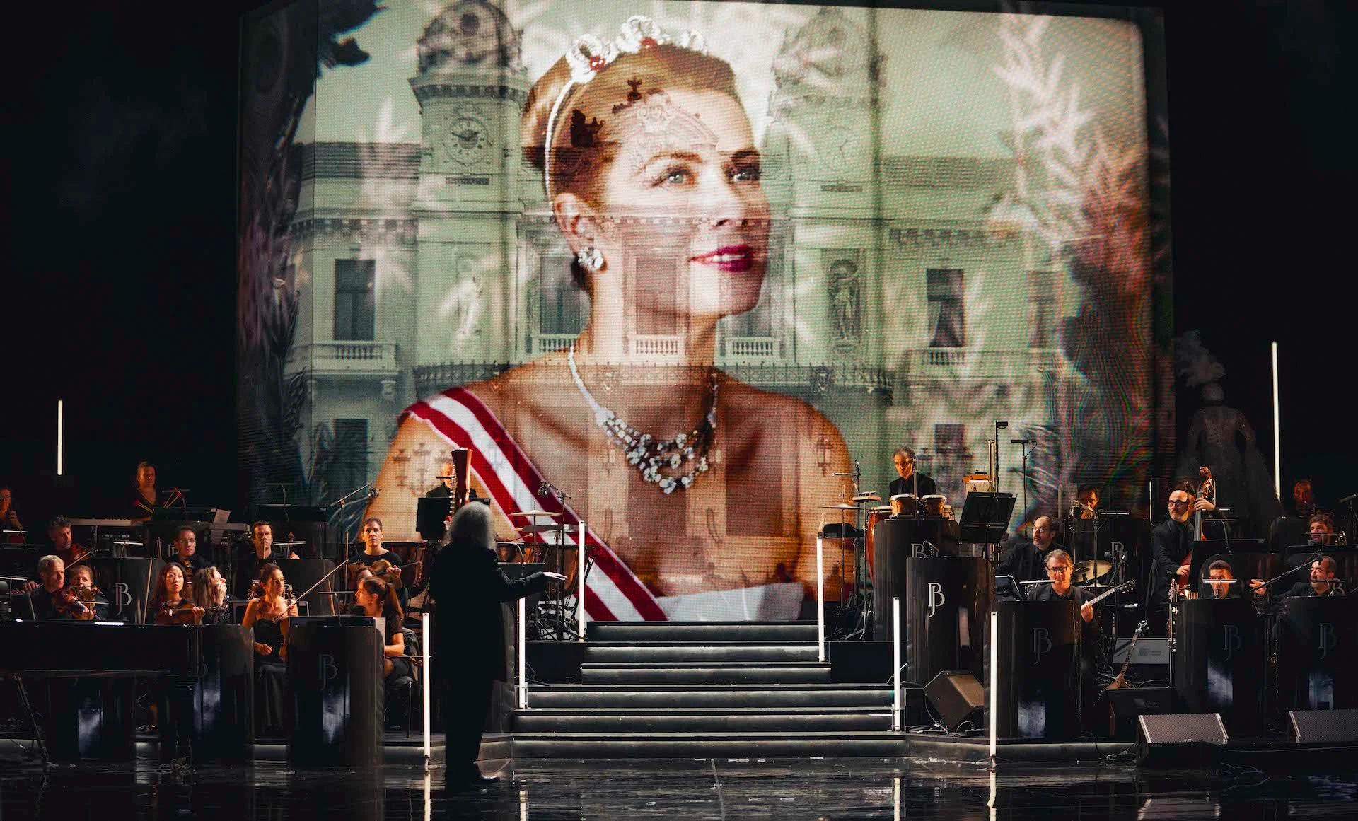 Ritratto proiettato di una donna con tiara e fascia sulla facciata di un palazzo, direttore e orchestra sotto – Bonsoir Monte-Carlo, Monte Carlo, Principato di Monaco, 2025 – scenografia
