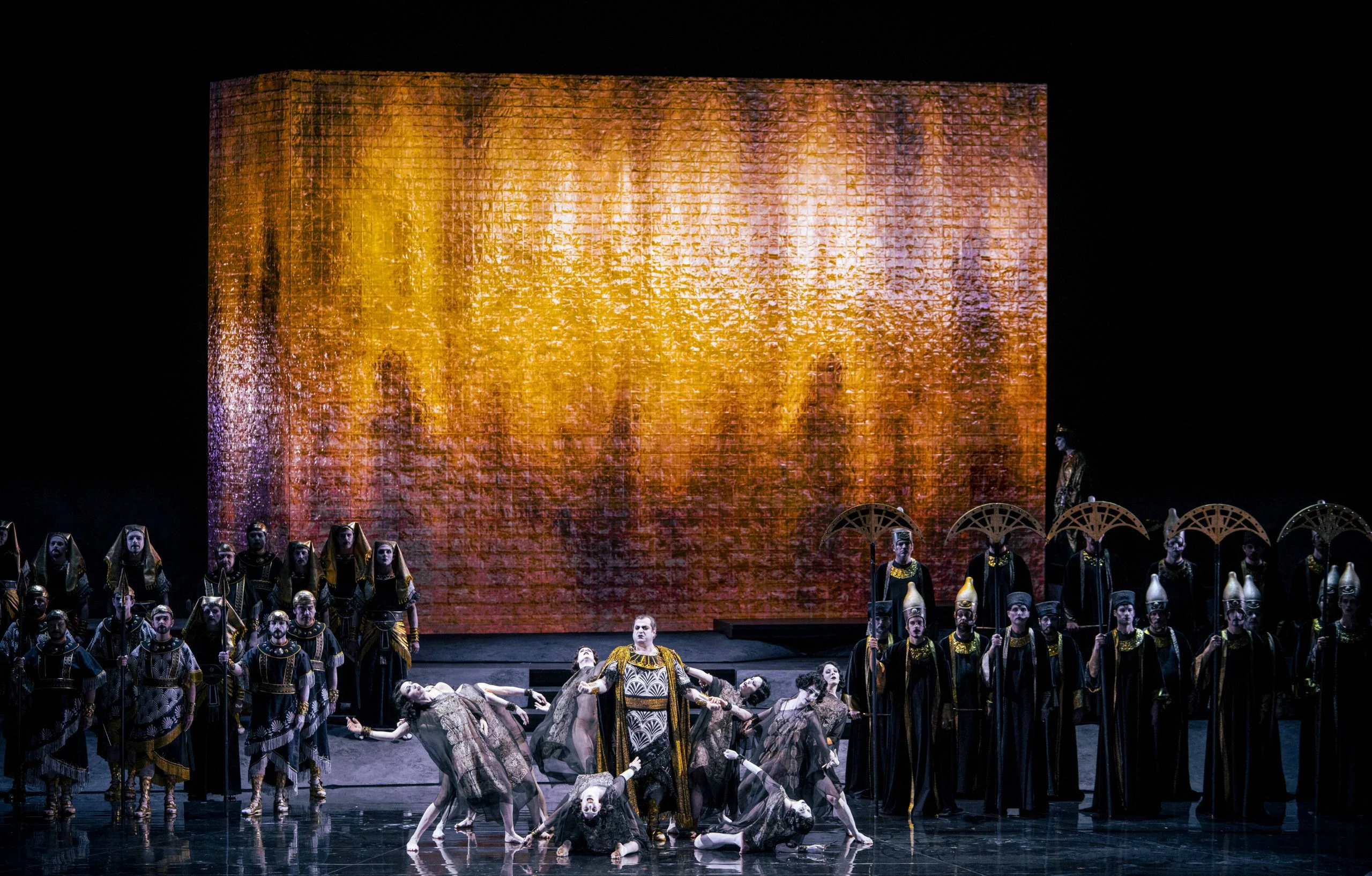 Ballerini attorno a un solista sul proscenio, coro ai lati e un blocco ambrato luminoso dietro – Aida all'Opéra de Monte-Carlo, Principato di Monaco, 2025 – scenografia d'opera