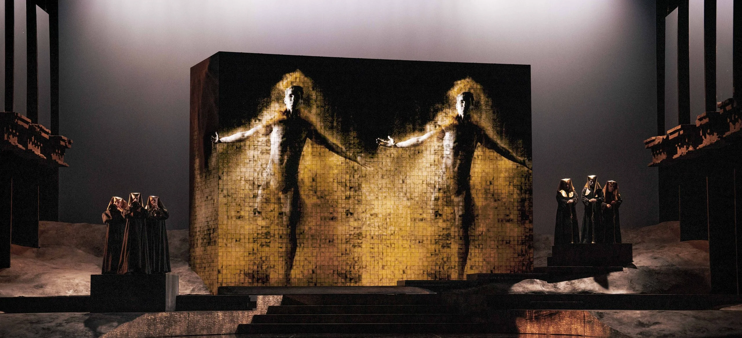 Blocco monumentale con proiezioni di due figure umane dorate, figure incappucciate ai lati – Aida all'Opéra de Monte-Carlo, Principato di Monaco, 2025 – scenografia d'opera