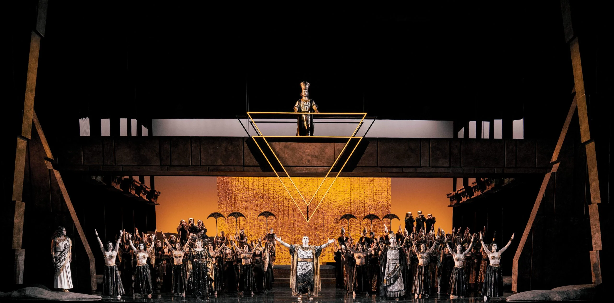 Scena trionfale: coro con le braccia alzate sotto un'alta galleria e un triangolo d'oro sospeso – Aida all'Opéra de Monte-Carlo, Principato di Monaco, 2025 – scenografia d'opera