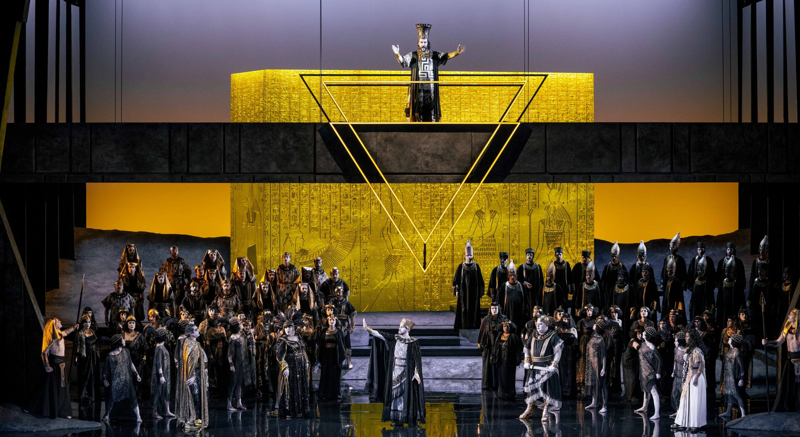 Coro dell'Aida in costumi egizi davanti a un blocco dorato di geroglifici, un sacerdote sulla galleria sopra un triangolo d'oro – Aida all'Opéra de Monte-Carlo, Principato di Monaco, 2025 – scenografia d'opera
