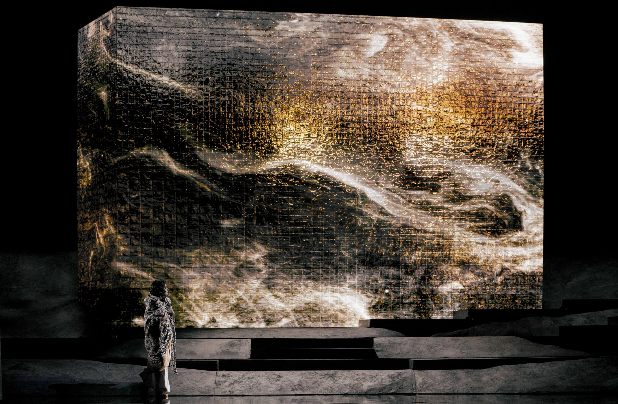 Figura solitaria in mantello bianco davanti a un blocco gigante con proiezioni di texture scure e dorate vorticose – Aida all'Opéra de Monte-Carlo, Principato di Monaco, 2025 – scenografia d'opera