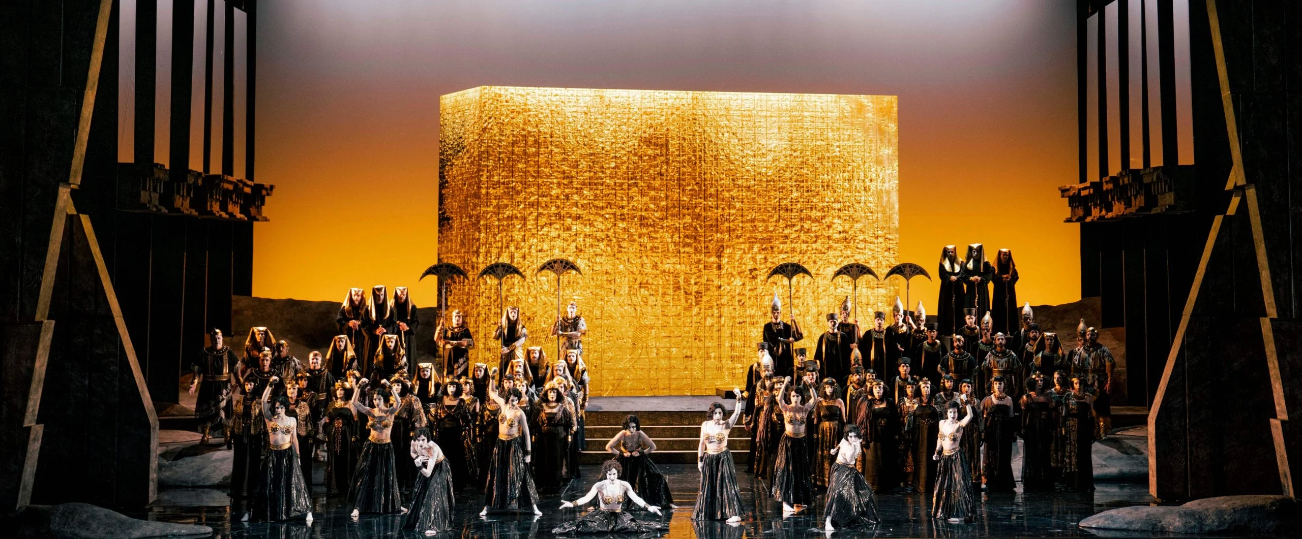 Vista d'insieme del blocco dorato contro un cielo ambrato, coro con parasole che riempie il palco – Aida all'Opéra de Monte-Carlo, Principato di Monaco, 2025 – scenografia d'opera