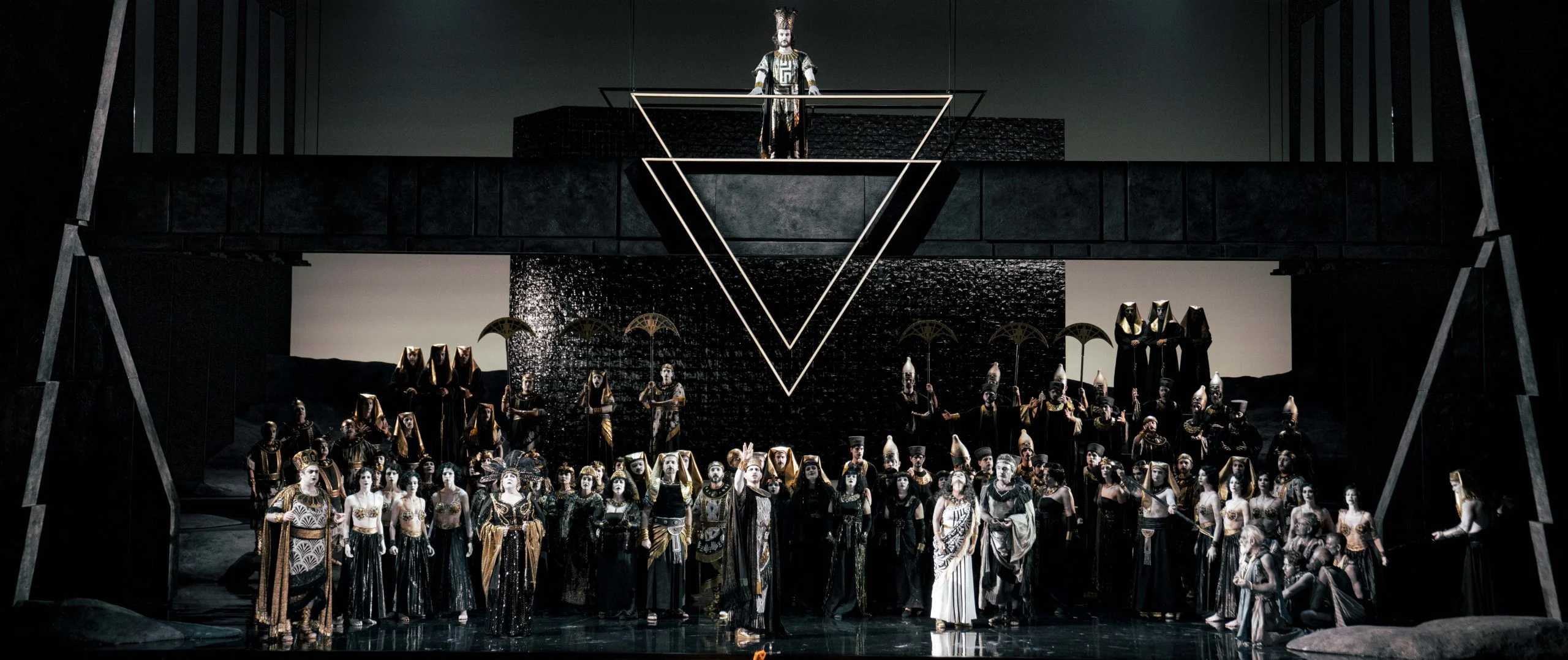 Scena in toni di grigio: coro affollato sotto un blocco scuro e una figura in piedi sopra un doppio triangolo – Aida all'Opéra de Monte-Carlo, Principato di Monaco, 2025 – scenografia d'opera