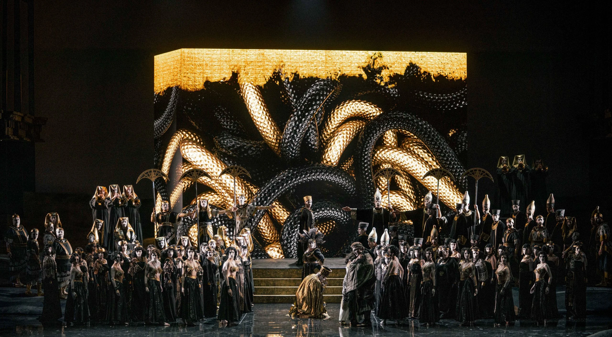 Blocco con proiezioni di serpenti attorcigliati oro e neri dietro il coro e una figura inginocchiata – Aida all'Opéra de Monte-Carlo, Principato di Monaco, 2025 – scenografia d'opera