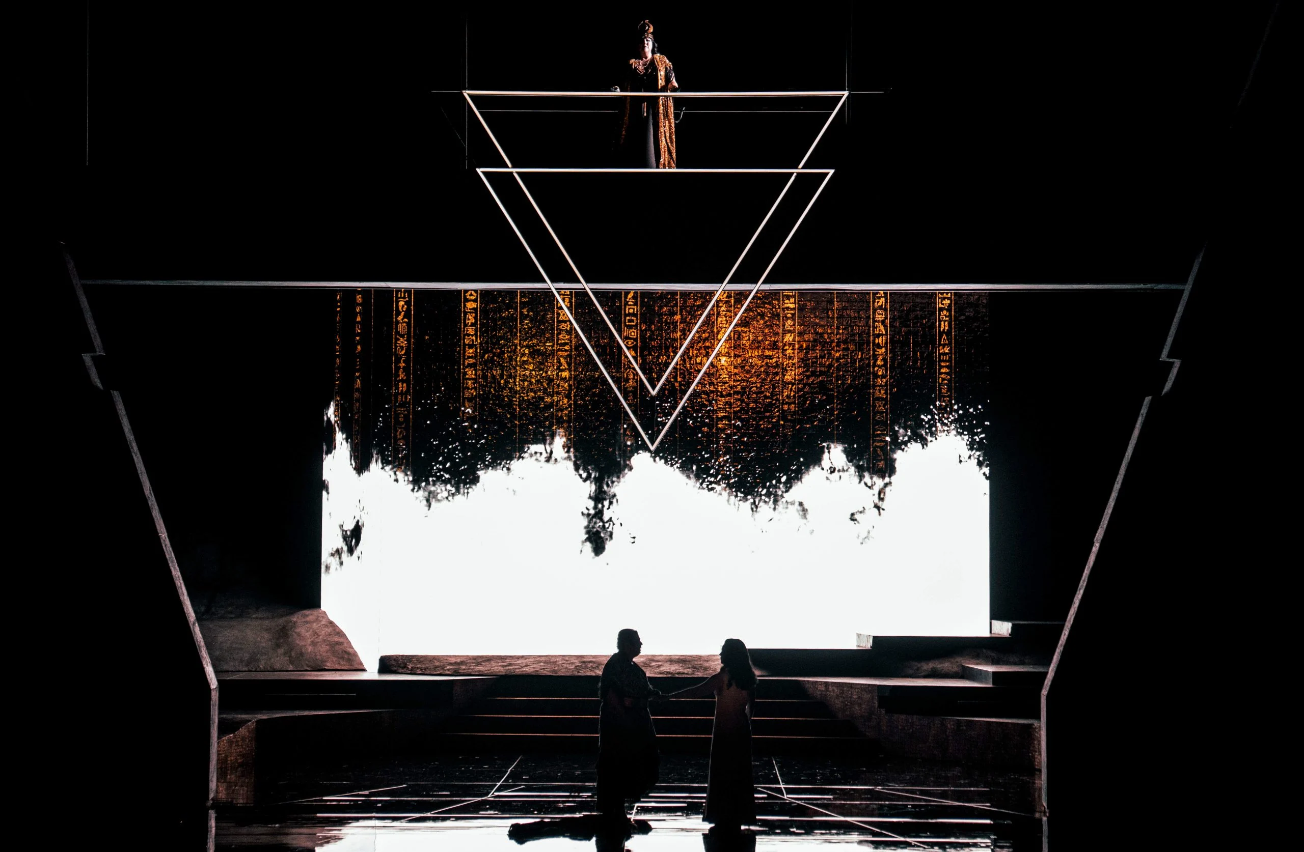 Due cantanti in silhouette contro uno schermo in luce bianca colante di geroglifici, un doppio triangolo sopra – Aida all'Opéra de Monte-Carlo, Principato di Monaco, 2025 – scenografia d'opera