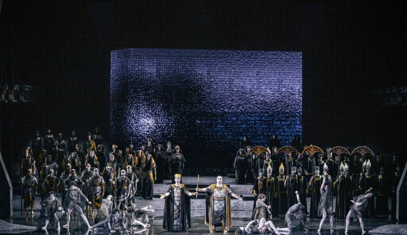 Coro e solisti su un palco riflettente davanti a un blocco monumentale illuminato da una fredda luce viola – Aida all'Opéra de Monte-Carlo, Principato di Monaco, 2025 – scenografia d'opera