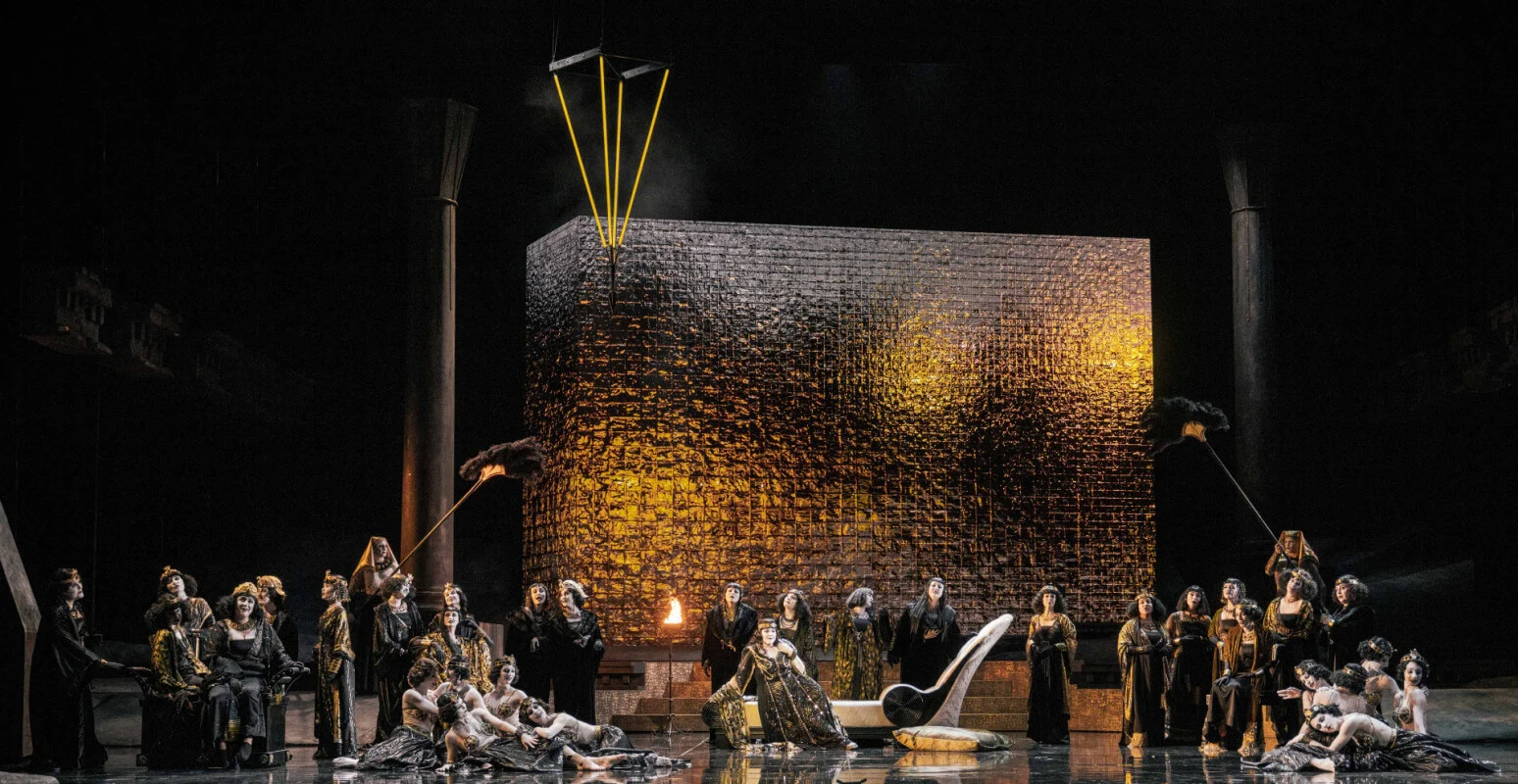Coro femminile disteso e seduto attorno a un grande blocco dorato materico, con ventagli di piume su aste – Aida all'Opéra de Monte-Carlo, Principato di Monaco, 2025 – scenografia d'opera