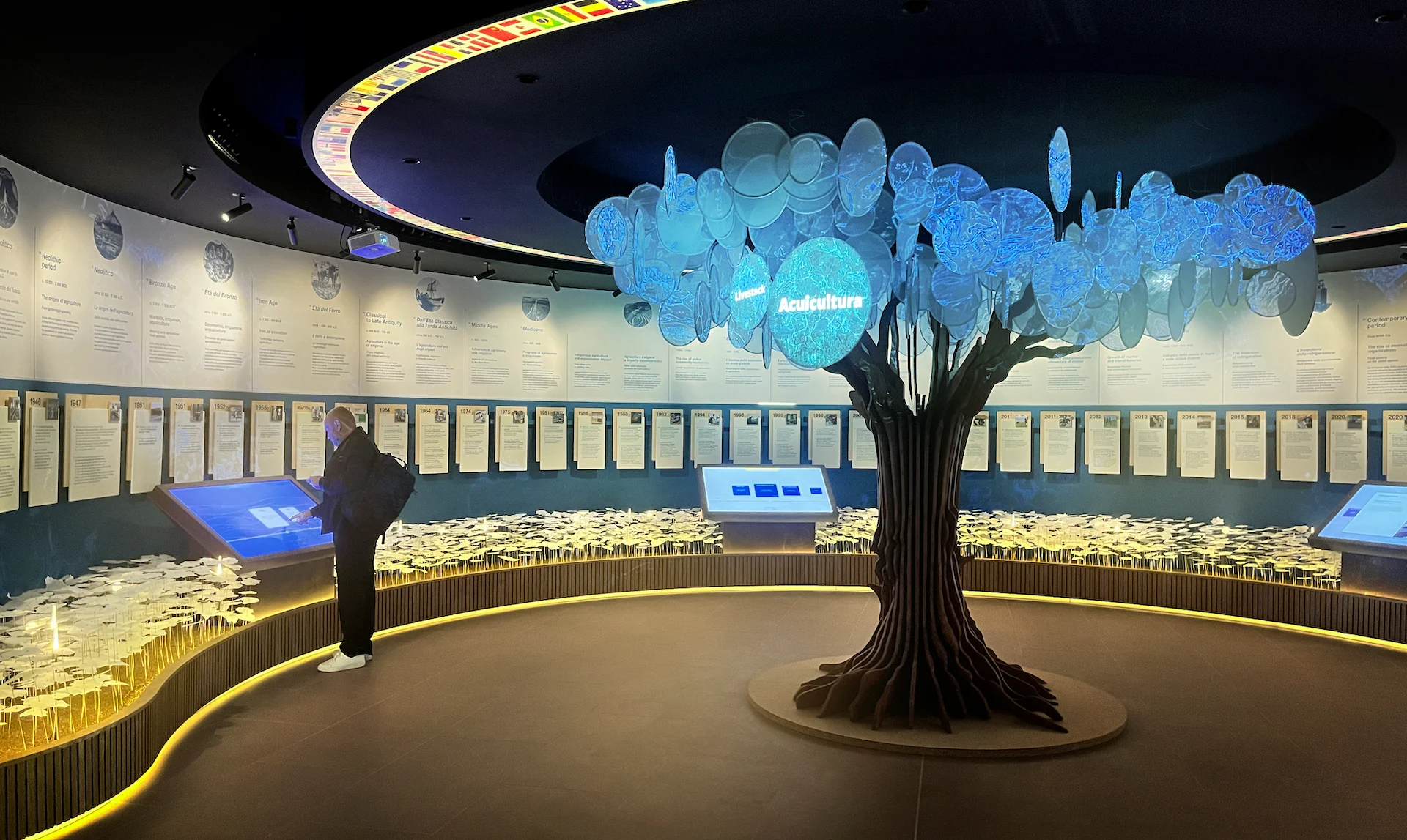 Sala museale circolare con un albero luminoso di dischi illuminati, schermi touch e pannelli a parete intorno – FAO MuNe Museo dell'alimentazione e dell'agricoltura, Roma, Italia, 2025 – allestimento espositivo