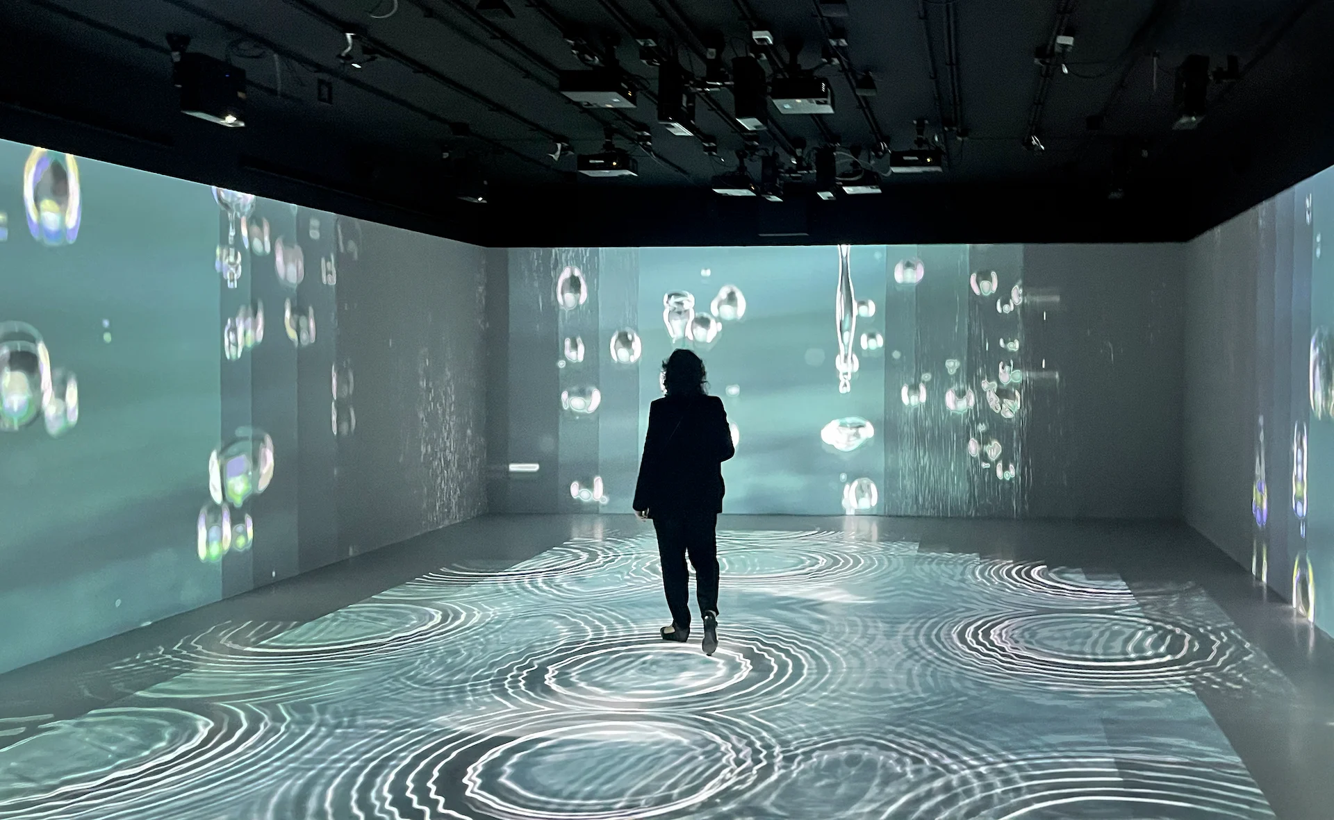 Visitatore che attraversa una sala immersiva con gocce d'acqua proiettate sulle pareti e increspature sul pavimento – FAO MuNe Museo dell'alimentazione e dell'agricoltura, Roma, Italia, 2025 – allestimento espositivo