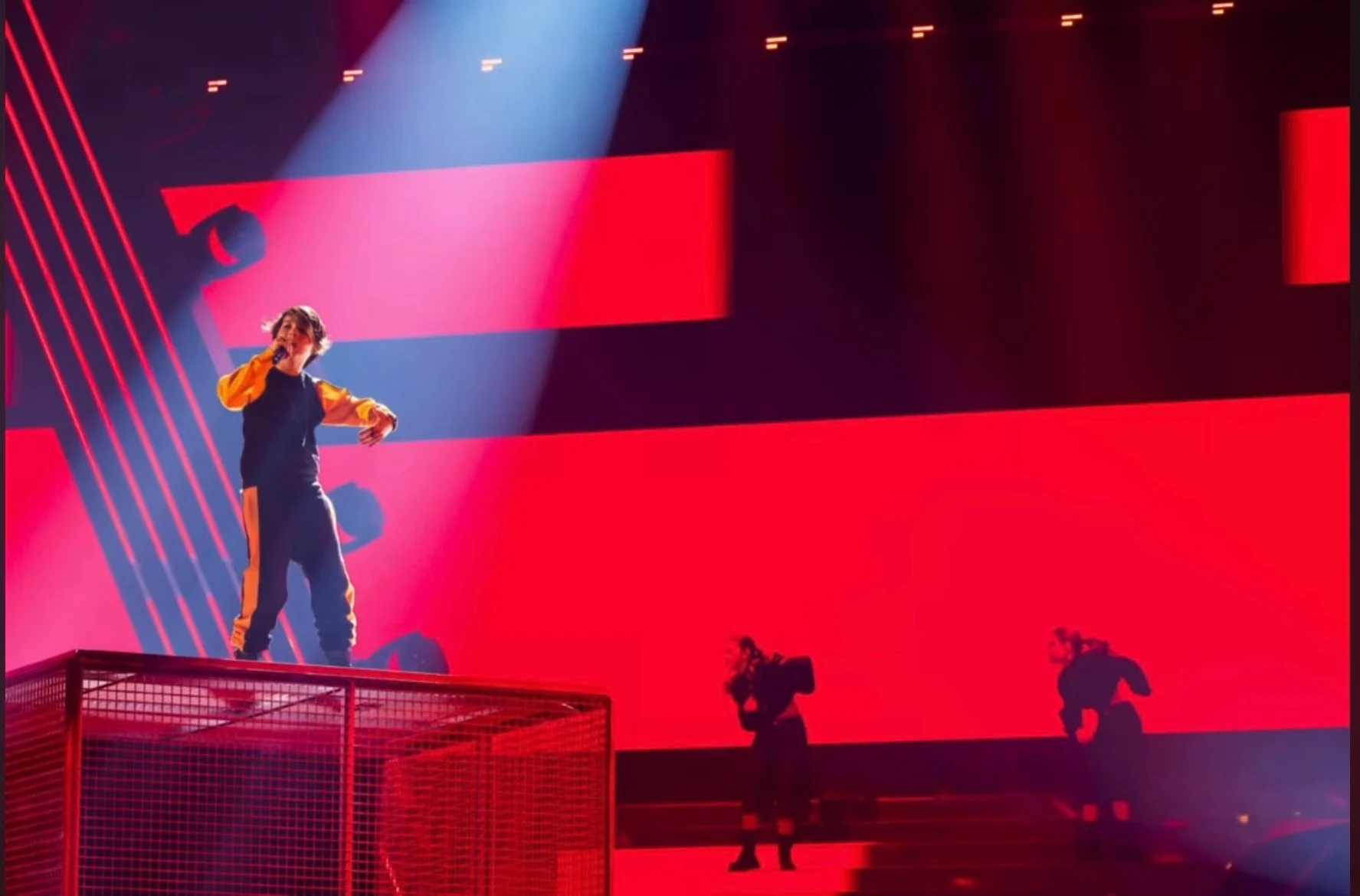 Cantante su una pedana a gabbia in rete contro un fondale LED rosso, con ballerini in silhouette sotto – Leonardo Giovannangeli al Junior Eurovision 2025, Tbilisi, Georgia – scenografia per concerti