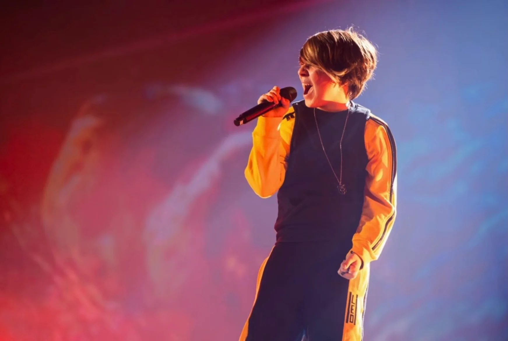 Primo piano del giovane cantante in tuta gialla e blu navy che canta davanti a uno schermo rosso e blu – Leonardo Giovannangeli al Junior Eurovision 2025, Tbilisi, Georgia – scenografia per concerti