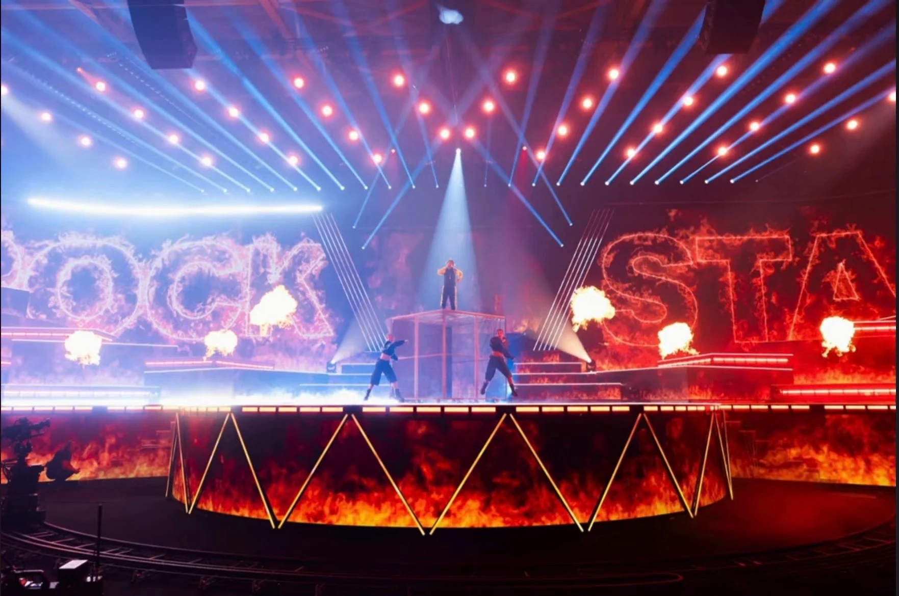 Vista d'insieme del palco di Rockstar: lettere LED infuocate, fiammate e una pedana bordata di luce sotto i fasci – Leonardo Giovannangeli al Junior Eurovision 2025, Tbilisi, Georgia – scenografia per concerti