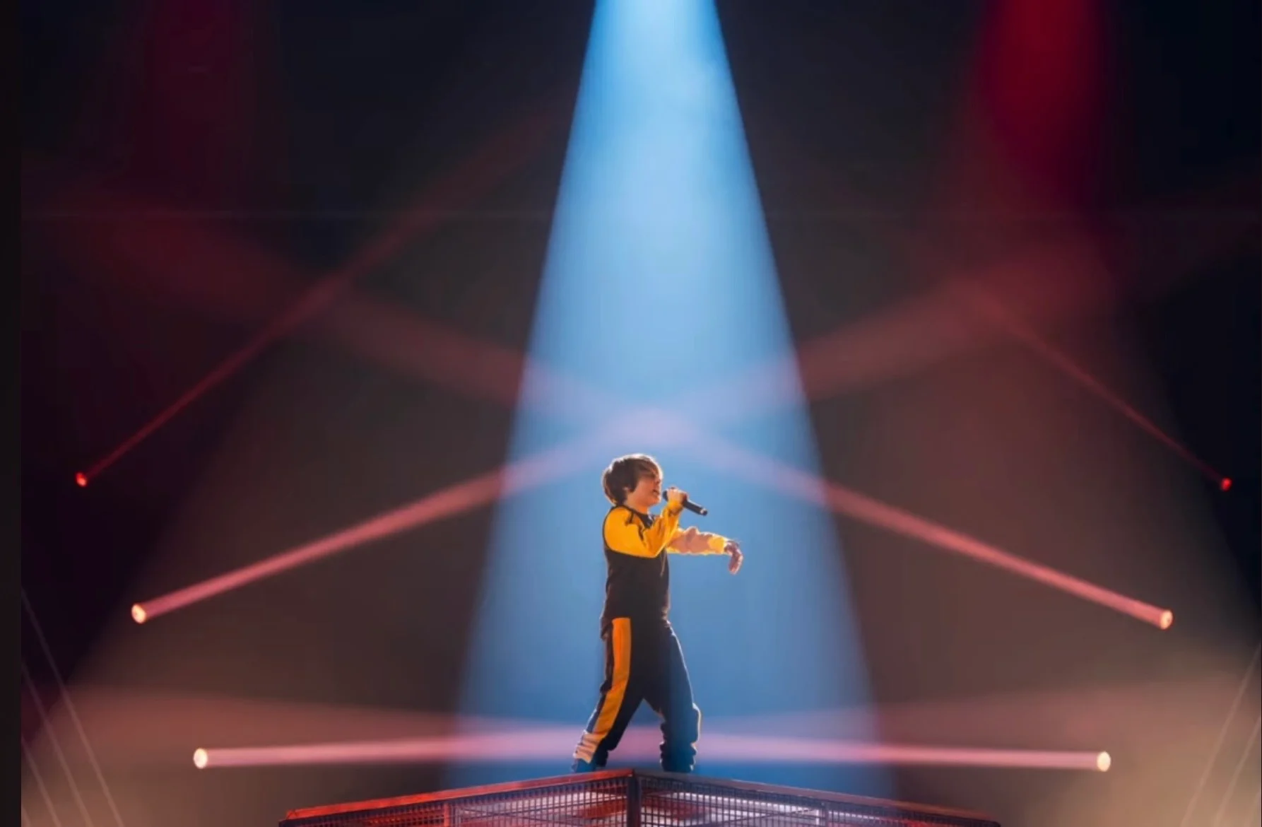 Cantante da solo sulla pedana a gabbia in un occhio di bue blu, attraversato da fasci di luce rossi – Leonardo Giovannangeli al Junior Eurovision 2025, Tbilisi, Georgia – scenografia per concerti