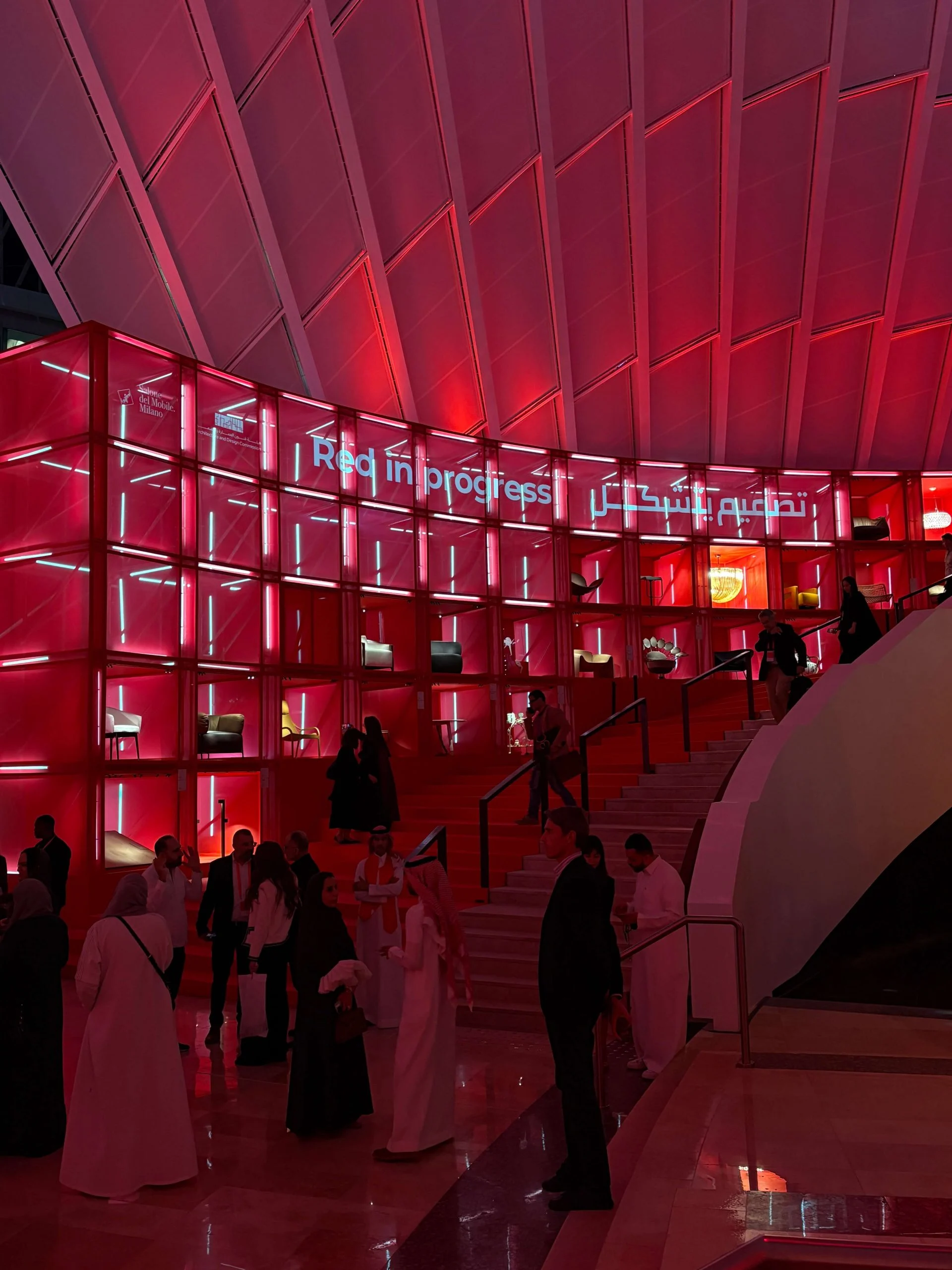 Folla serale ai piedi della parete espositiva rossa illuminata e della scala – Red in Progress: Salone del Mobile Meets Riyadh, Arabia Saudita, 2025 – allestimento espositivo