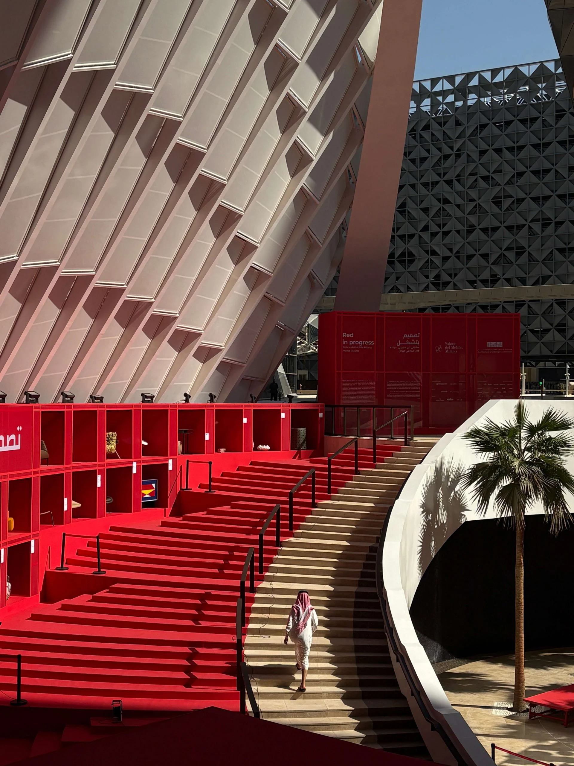Visitatore che scende una scala accanto a scaffali espositivi rossi e gradoni rossi sotto una copertura bianca nervata – Red in Progress: Salone del Mobile Meets Riyadh, Arabia Saudita, 2025 – allestimento espositivo