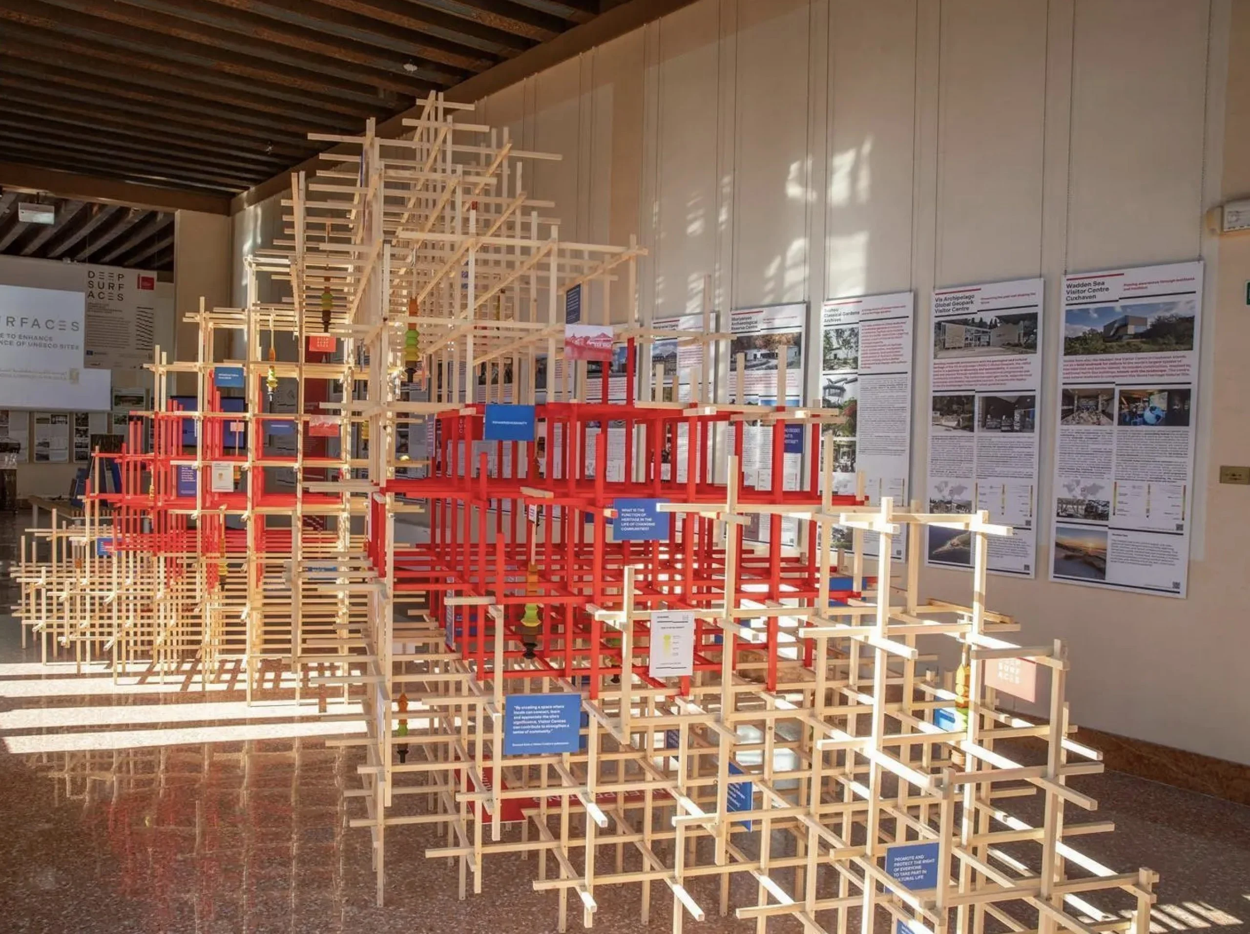 Installazione a graticcio in legno con telai rossi e naturali in una sala soleggiata, pannelli dei progetti lungo la parete – Deep Surfaces a Palazzo Zorzi, Venezia, Italia, 2025 – allestimento espositivo