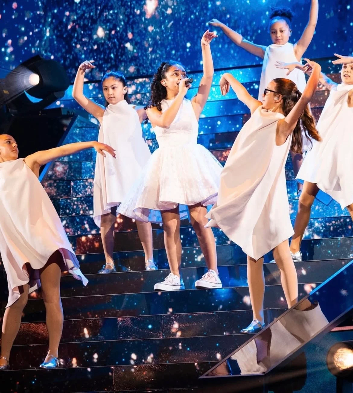 Cantante e ballerine in abiti bianchi su gradini illuminati contro un fondale LED blu stellato – Eliza Borg al Junior Eurovision 2025, Tbilisi, Georgia – scenografia per concerti