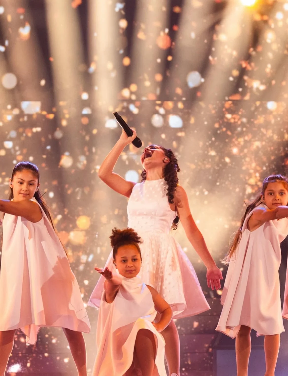 La giovane cantante maltese in abito bianco tra i ballerini, sotto fasci di luce dorata e scintillii – Eliza Borg al Junior Eurovision 2025, Tbilisi, Georgia – scenografia per concerti