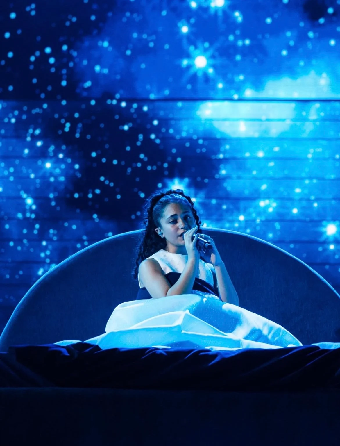 Cantante seduta su un letto a forma di falce di luna sotto un cielo notturno stellato sullo schermo LED – Eliza Borg al Junior Eurovision 2025, Tbilisi, Georgia – scenografia per concerti
