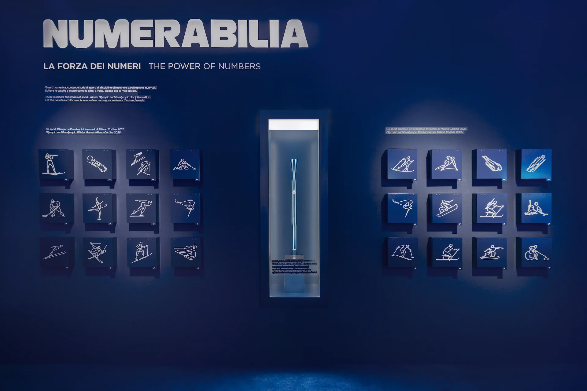 Parete Numerabilia: pittogrammi degli sport olimpici su piastrelle blu ai lati di una vetrina illuminata – Casa Esselunga a Milano Cortina 2026, Italia – allestimento espositivo