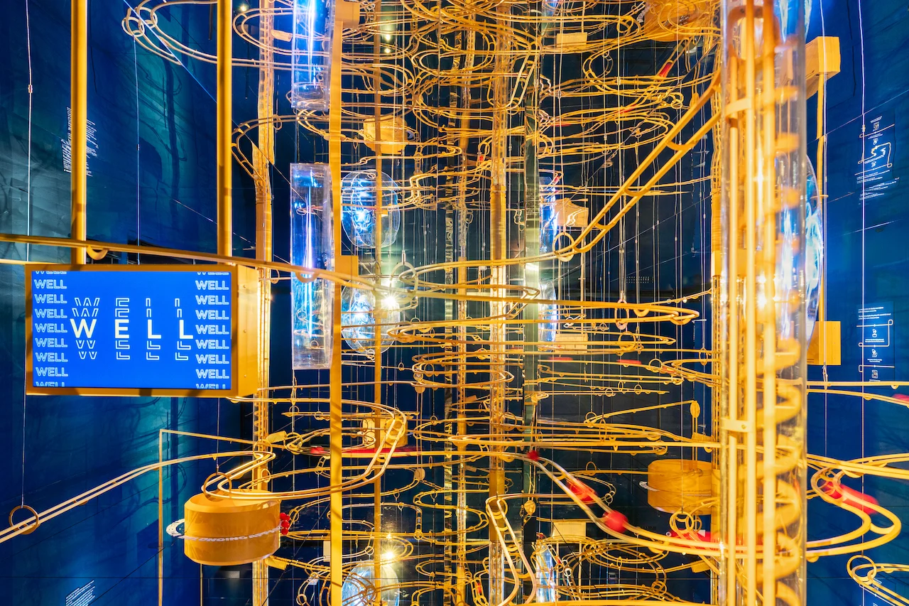 Fitto intreccio di piste per biglie dorate con uno schermo blu con la scritta WELL, in una stanza blu a specchi – Casa Esselunga a Milano Cortina 2026, Italia – allestimento espositivo