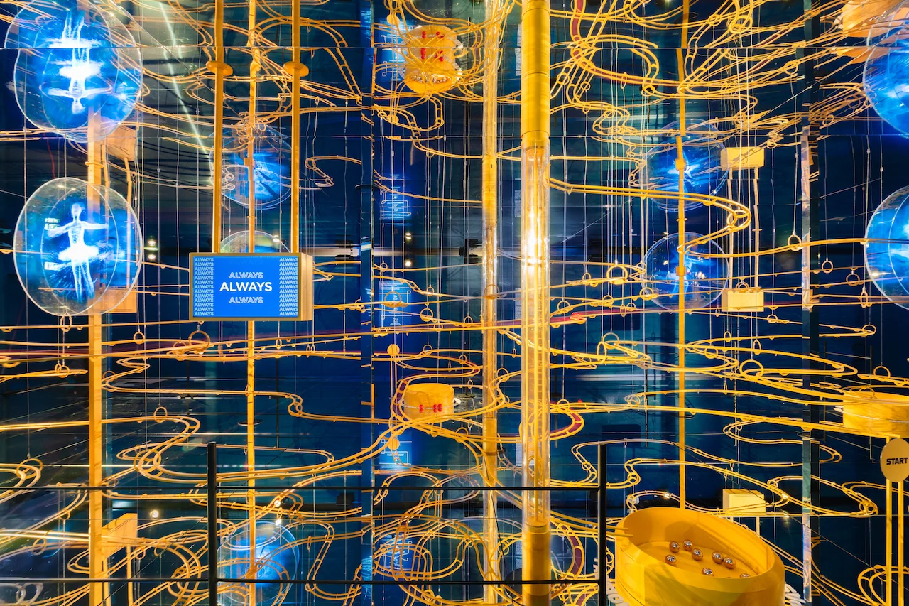 Vista d'insieme dell'installazione con le piste per biglie: binari dorati, ologrammi luminosi di atleti e la scritta ALWAYS – Casa Esselunga a Milano Cortina 2026, Italia – allestimento espositivo