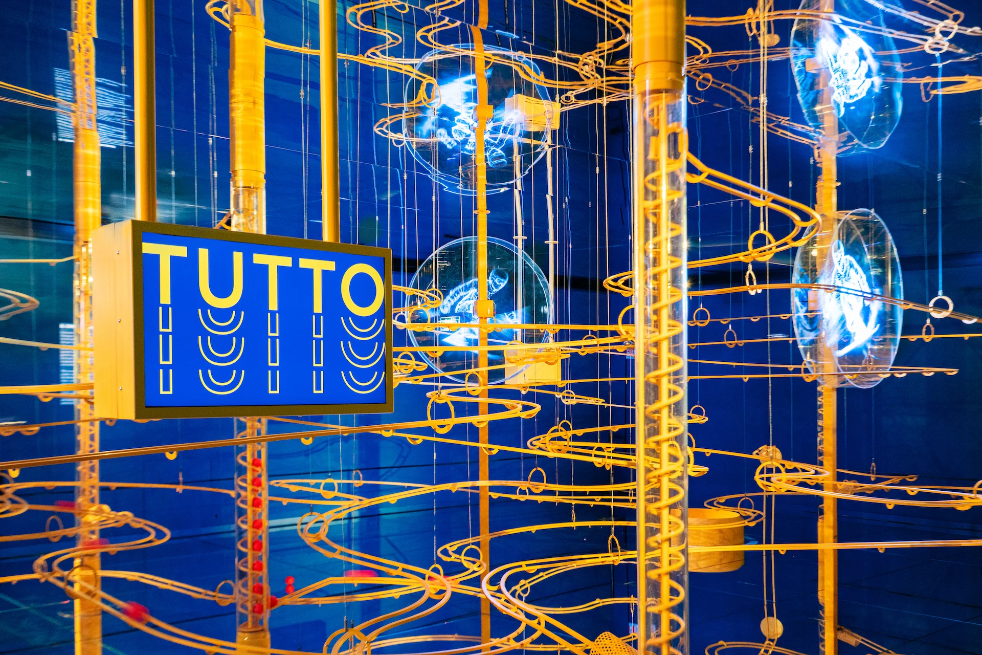 Dettaglio di piste dorate a spirale e dischi olografici accanto a un'insegna blu con la scritta TUTTO – Casa Esselunga a Milano Cortina 2026, Italia – allestimento espositivo