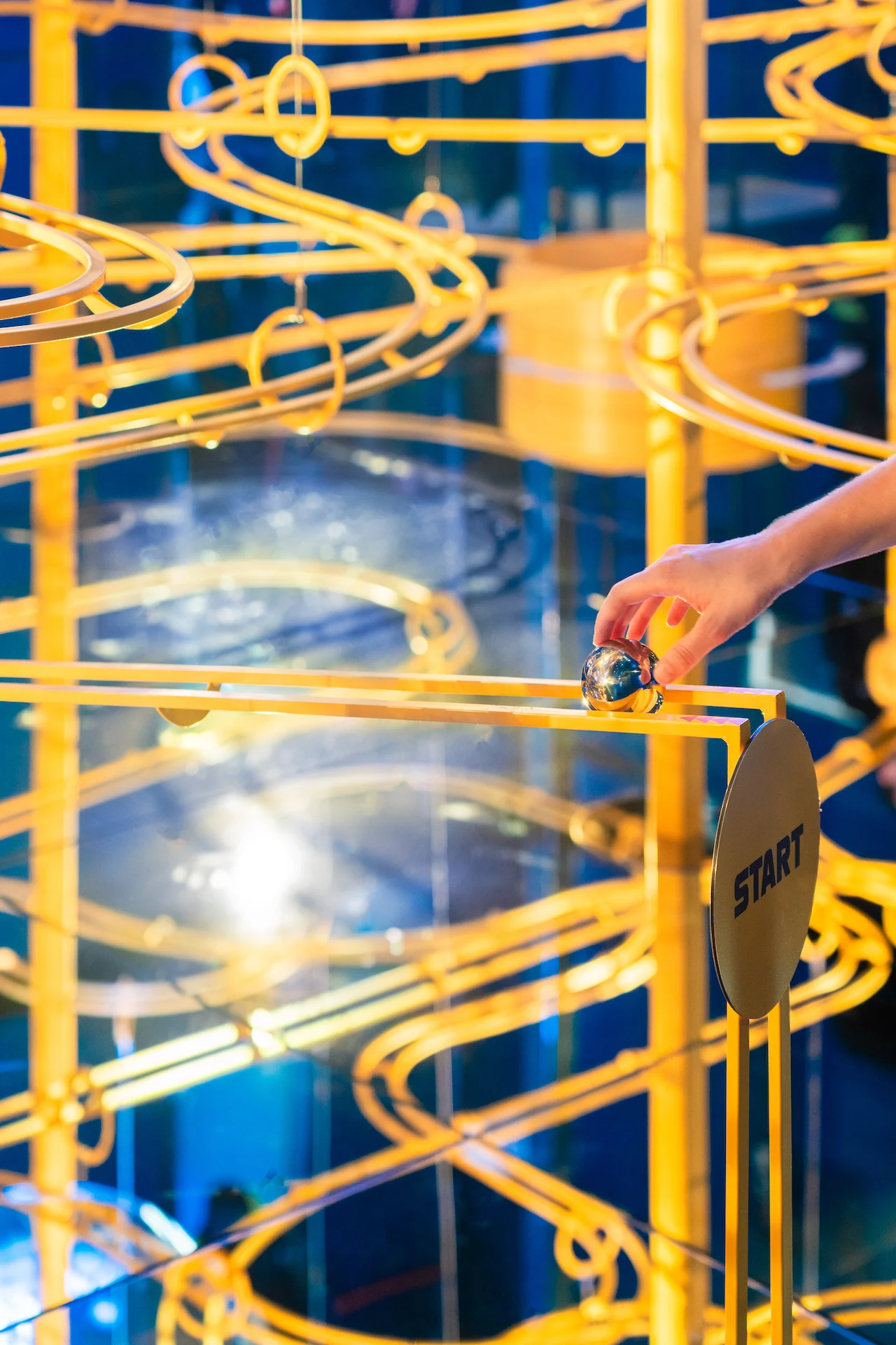 Una mano posa una sfera d'acciaio all'inizio di una pista per biglie dorata in una stanza blu a specchi – Casa Esselunga a Milano Cortina 2026, Italia – allestimento espositivo