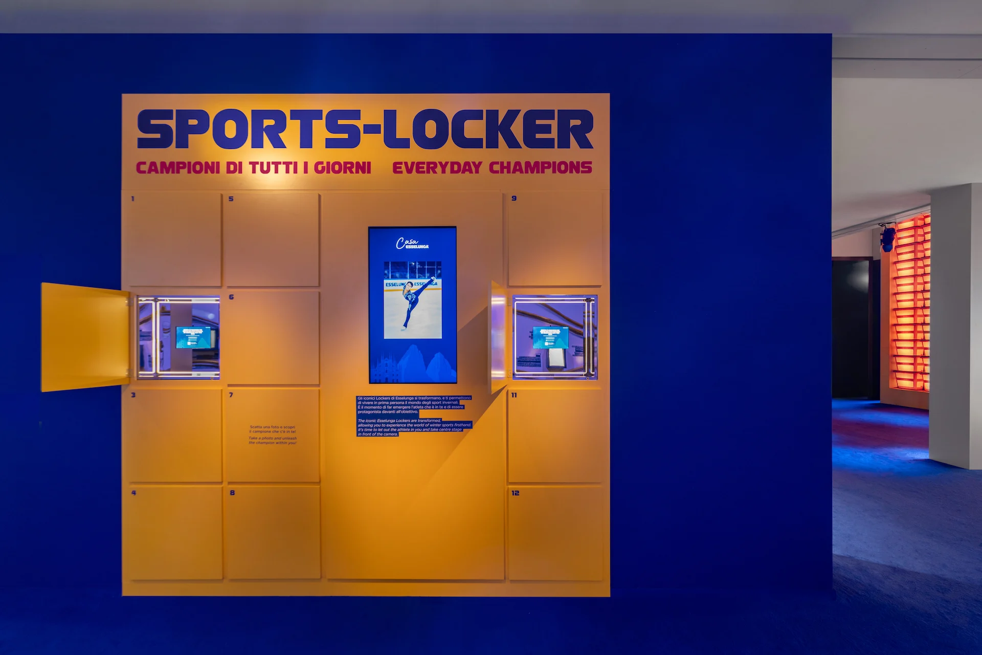 Parete gialla Sports-Locker con ante aperte che mostrano piccoli schermi, in una stanza blu intenso – Casa Esselunga a Milano Cortina 2026, Italia – allestimento espositivo