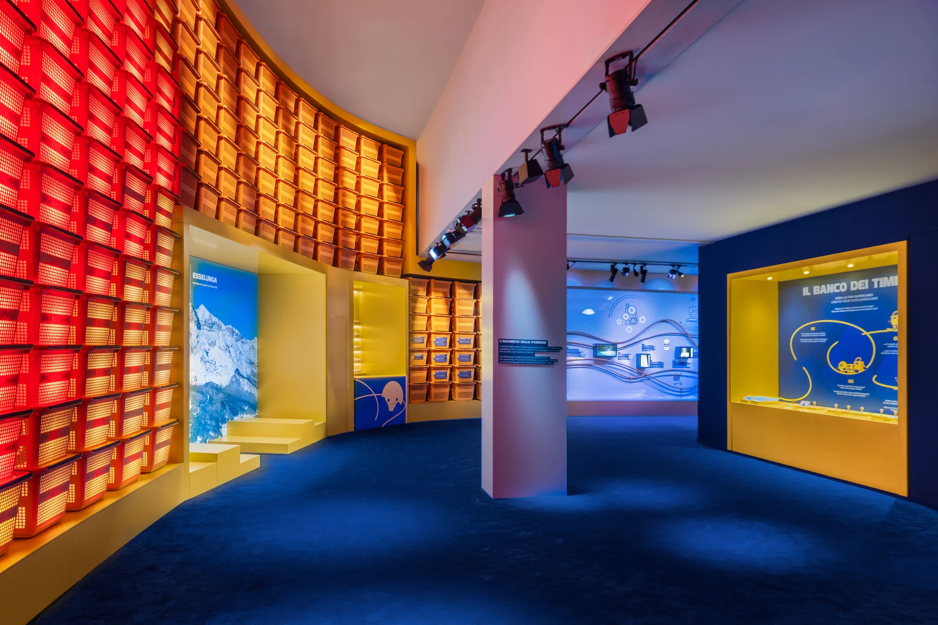 Sala espositiva con una parete curva di cestini della spesa rossi, l'immagine di una montagna e nicchie espositive gialle – Casa Esselunga a Milano Cortina 2026, Italia – allestimento espositivo
