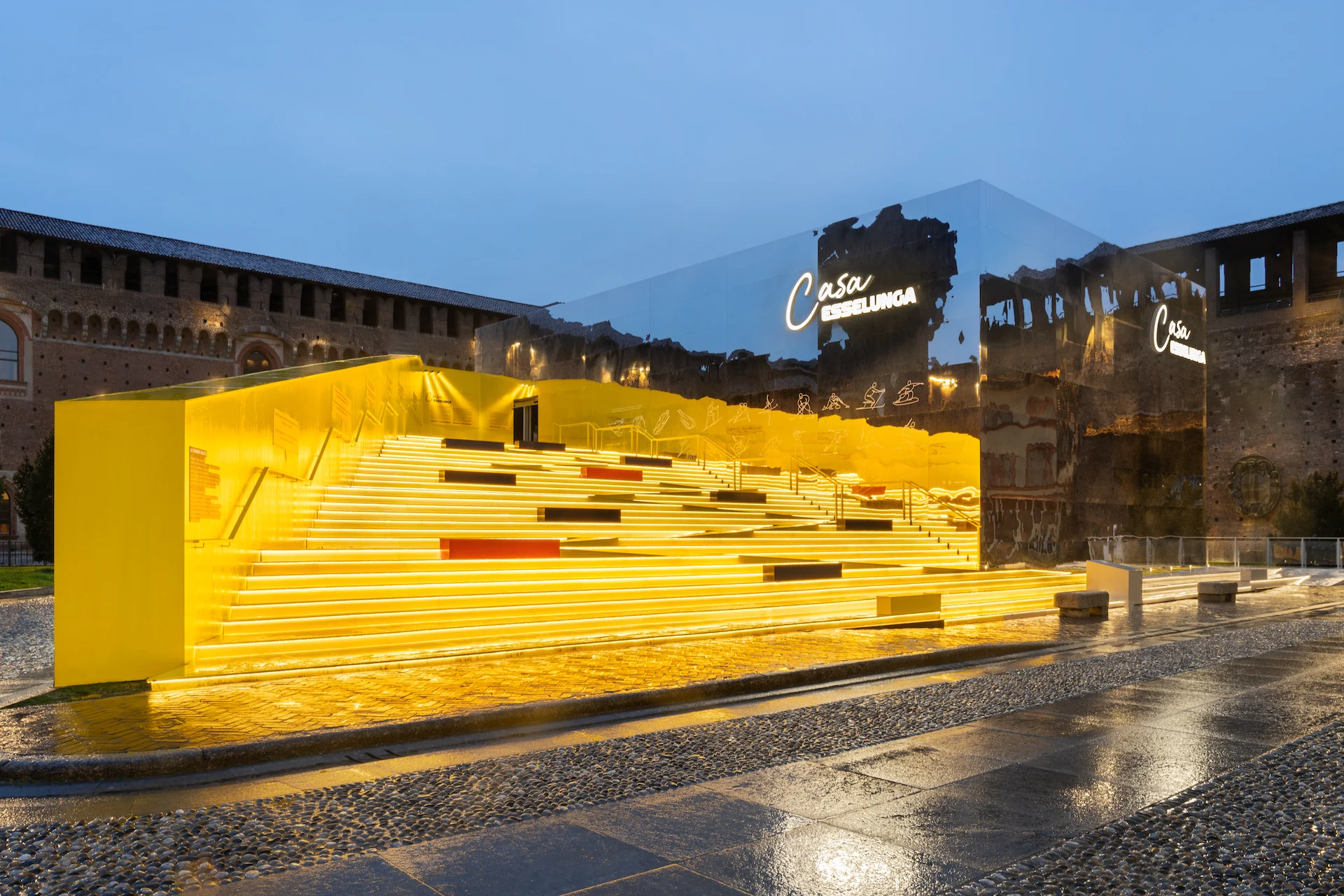 Casa Esselunga al crepuscolo: struttura gialla a gradoni illuminata e padiglione a specchio nel cortile del Castello Sforzesco – Casa Esselunga a Milano Cortina 2026, Italia – allestimento espositivo