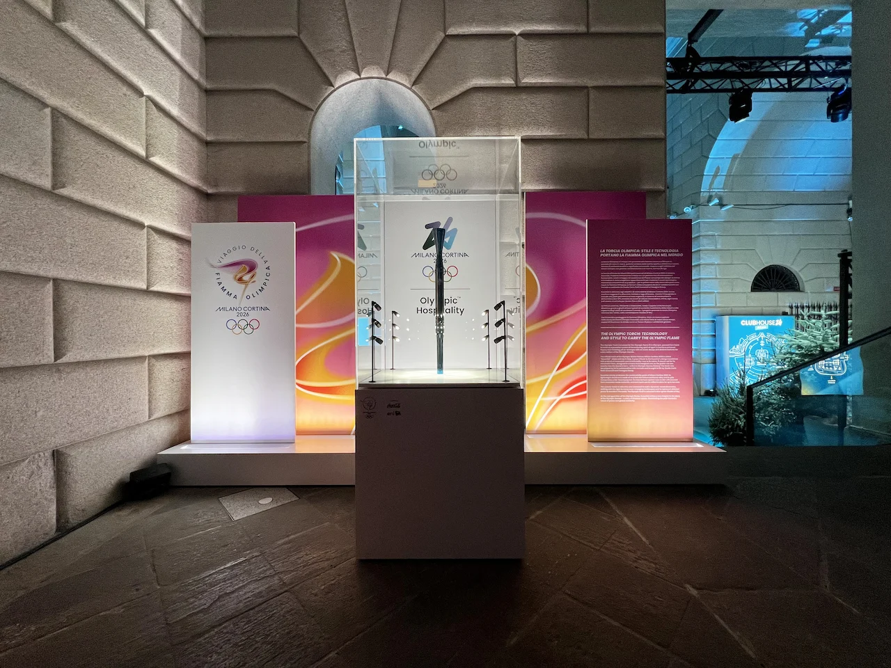 Teca con una torcia olimpica davanti a pannelli colorati di Milano Cortina 2026, contro una parete di pietra – Clubhouse 26 Milano, Italia, 2026 – hospitality design