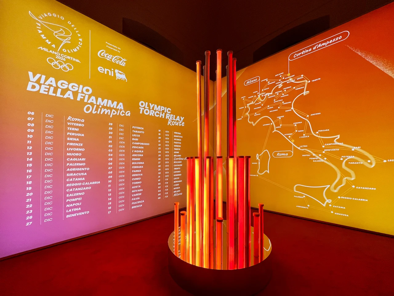 Sala della staffetta della fiamma: scultura di tubi luminosi rossi e arancioni davanti a pareti con la mappa del percorso in Italia – Clubhouse 26 Milano, Italia, 2026 – hospitality design