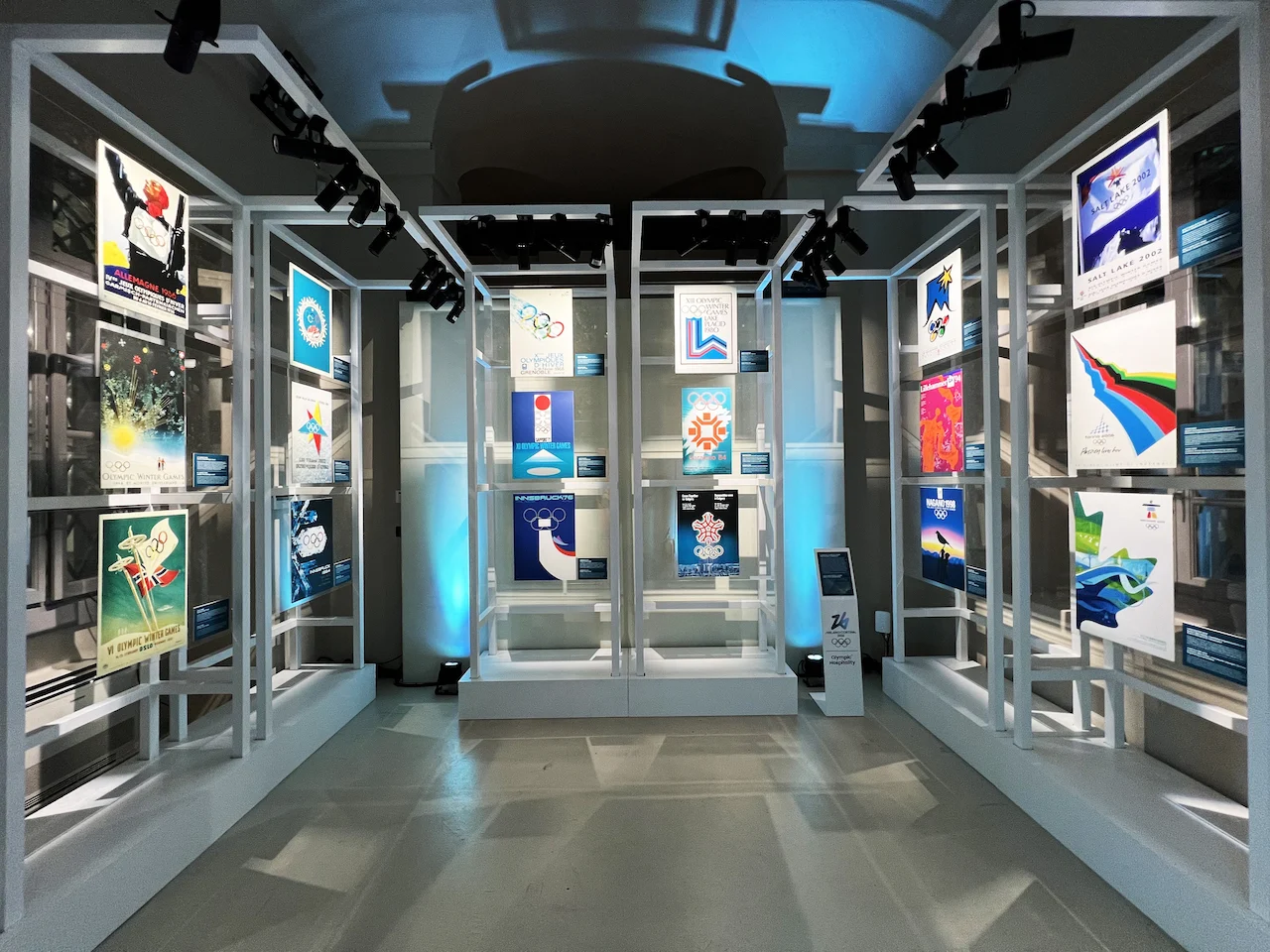 Strutture a telaio bianche con manifesti storici delle Olimpiadi invernali sotto i riflettori e luce blu – Clubhouse 26 Milano, Italia, 2026 – hospitality design