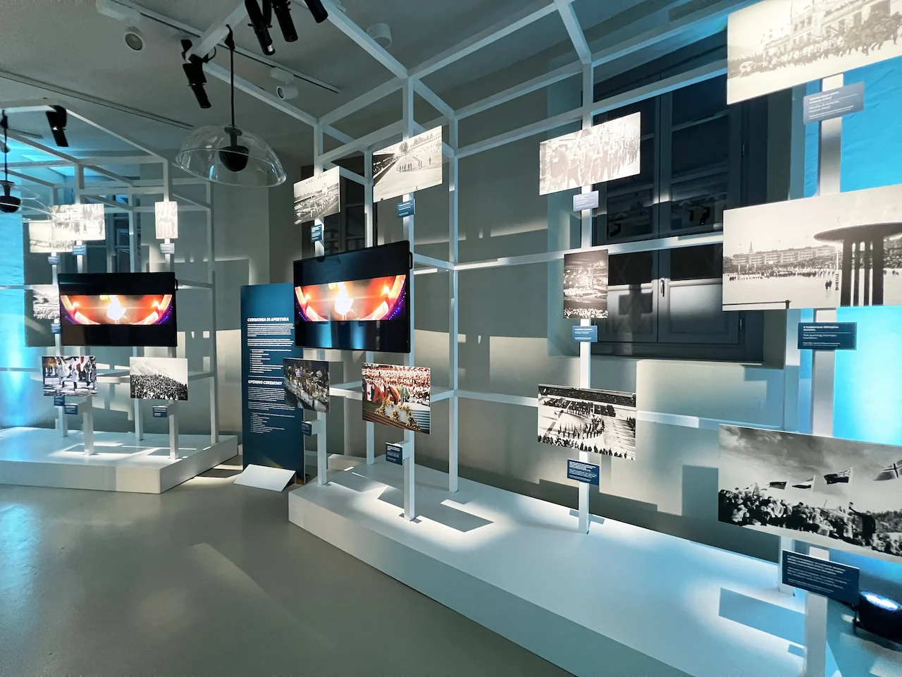 Fotografie olimpiche d'archivio e schermi montati su telai bianchi aperti, in una fredda luce blu – Clubhouse 26 Milano, Italia, 2026 – hospitality design
