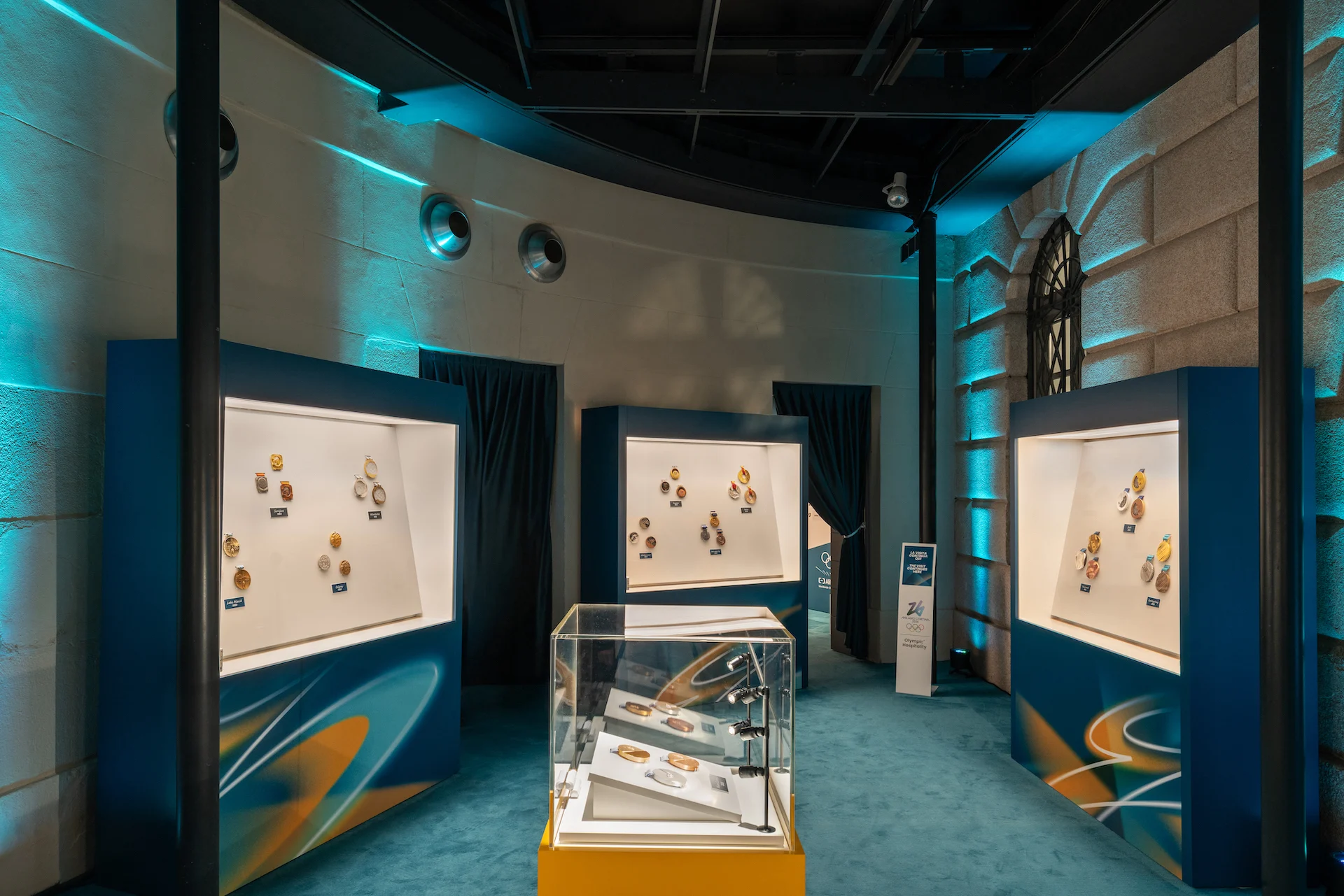 Sala espositiva curva con nicchie illuminate di medaglie olimpiche e una teca centrale, sotto una luce verde acqua – Clubhouse 26 Milano, Italia, 2026 – hospitality design
