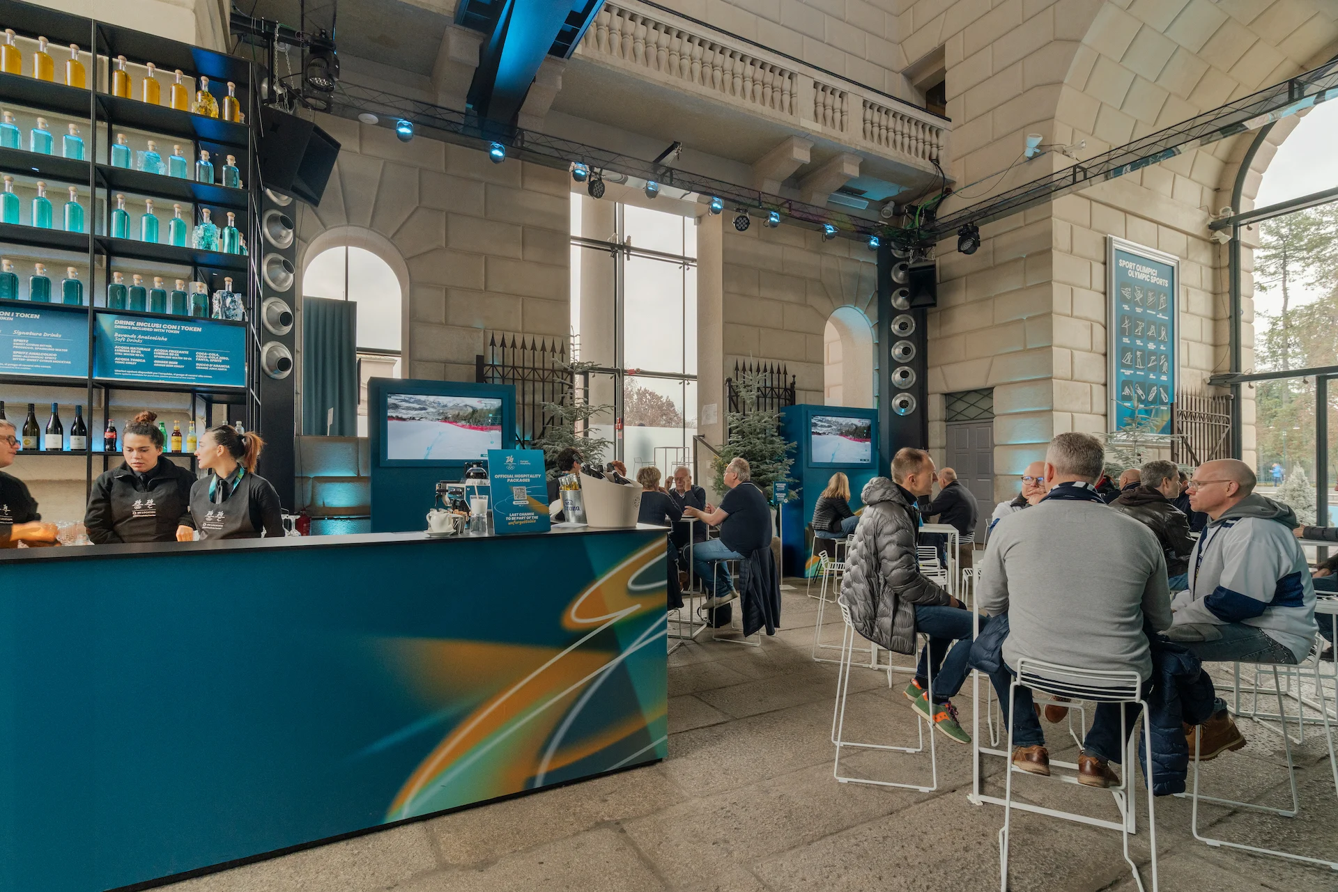 Lounge hospitality con un bancone curvo blu, tavoli alti e ospiti, in una sala di pietra con grandi finestre – Clubhouse 26 Milano, Italia, 2026 – hospitality design